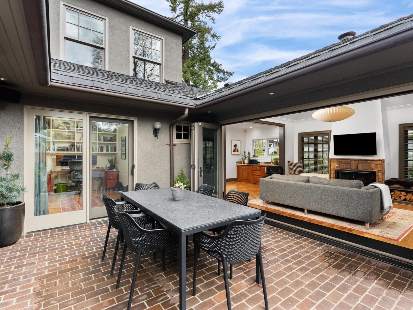 A modern home in Portland, Oregon, with a spacious patio featuring a dining table and chairs. Large sliding doors open to a cozy interior with a sofa, coffee table, and fireplace. The surrounding greenery and clear blue sky create the perfect setting for anyone seeking quality Portland real estate.