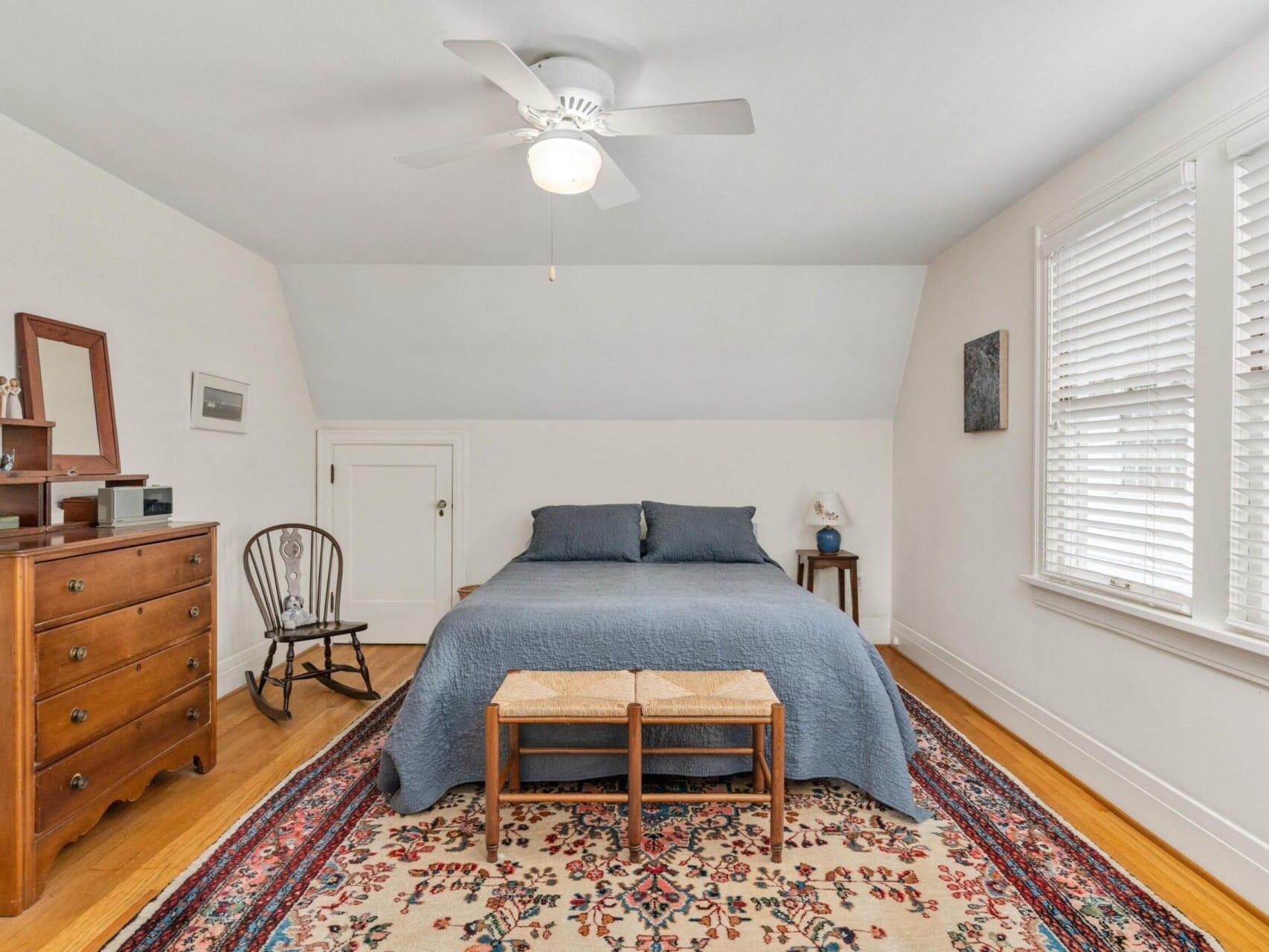 A cozy Portland, Oregon bedroom features a blue bedspread on a double bed, flanked by a wooden dresser and chair. A colorful rug graces the wooden floor, with a ceiling fan overhead. Small windows with blinds and framed pictures adorn the white walls, adding charm to this inviting space.