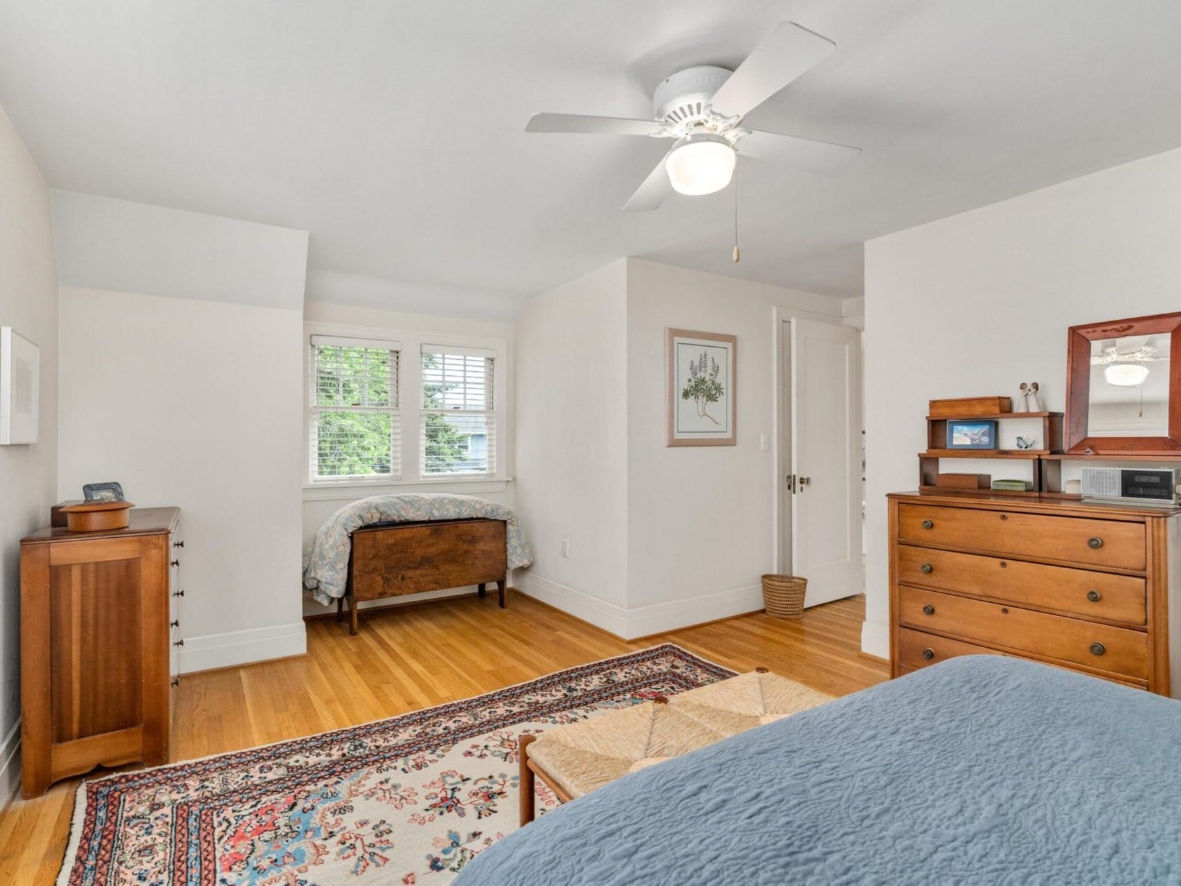 A bright bedroom in Portland, Oregon, boasts a blue bedspread, wooden dresser, and ceiling fan. A twin bed nestles in an alcove by the windows. The room features wooden floors, a patterned rug, and framed artwork on the walls. Natural light fills the inviting space.