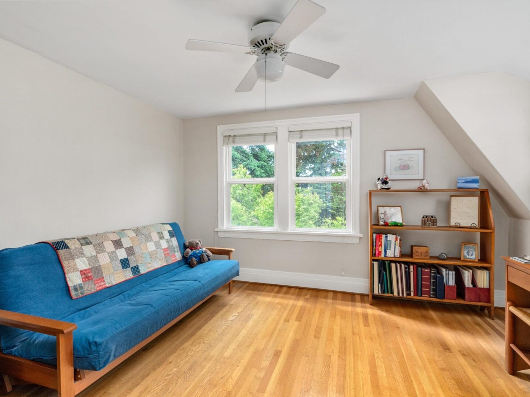 A bright room in Portland, Oregon, with a blue futon, a ceiling fan, and a wooden bookshelf filled with books and decor. Sunlight streams through the window, illuminating the wooden dresser on the right. The space boasts wooden flooring and pristine white walls.