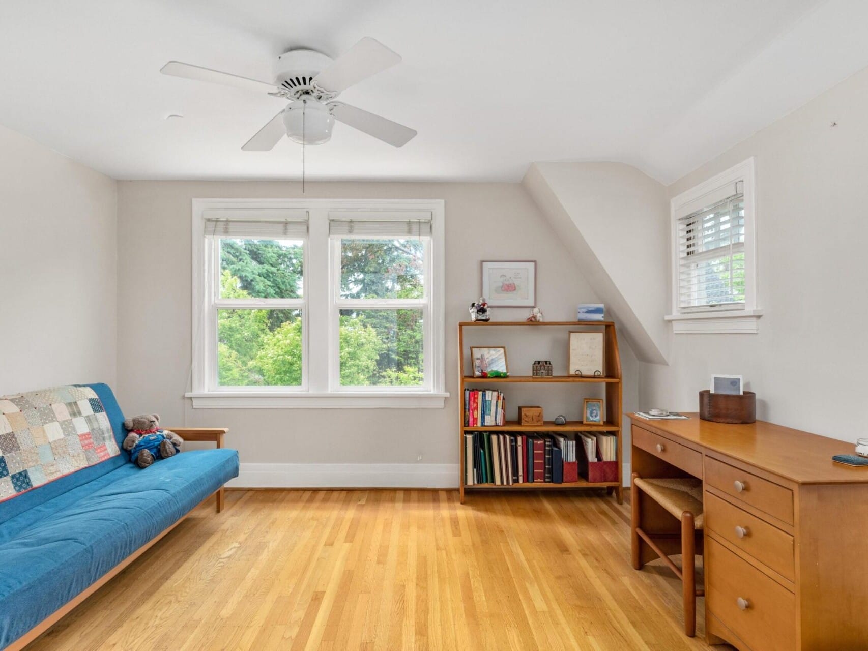 A cozy Portland Oregon room with a blue sofa and a colorful patchwork quilt. A small teddy bear sits on the sofa. There is a wooden bookshelf with books and decor items, and a desk with drawers. Two large windows and a ceiling fan brighten the space.