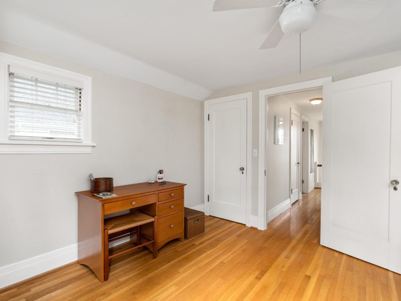 A simple bedroom in Portland, Oregon, showcases light gray walls and a wooden desk with drawers. A ceiling fan circulates air above the hardwood floor. To the side, a small window with blinds adds light, and two doors open to a hallway in the background.