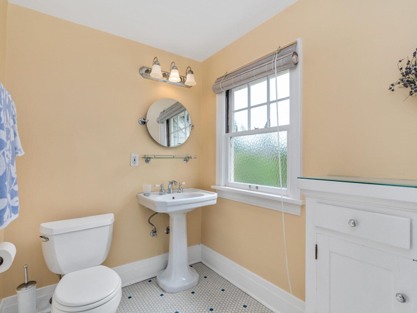 A cozy Portland, Oregon bathroom with yellow walls features a pedestal sink, a circular mirror, and a white toilet. A window with a blind sits above the cabinet. A towel hangs on the wall, while a floral wreath offers charm. A three-bulb light fixture illuminates above the mirror.