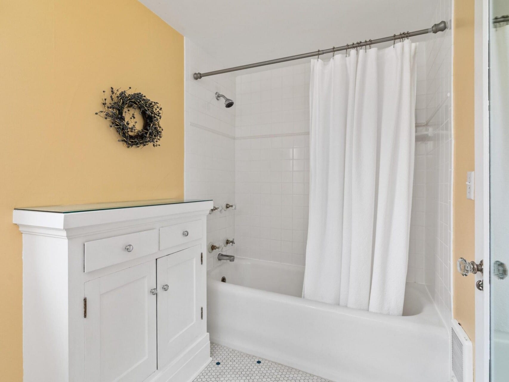 A cozy Portland, Oregon bathroom features a white bathtub with shower curtain, tiled walls, and a white vanity offering ample storage. The walls are painted warm yellow with a decorative wreath above the vanity, while the floor boasts a small hexagonal tile pattern.