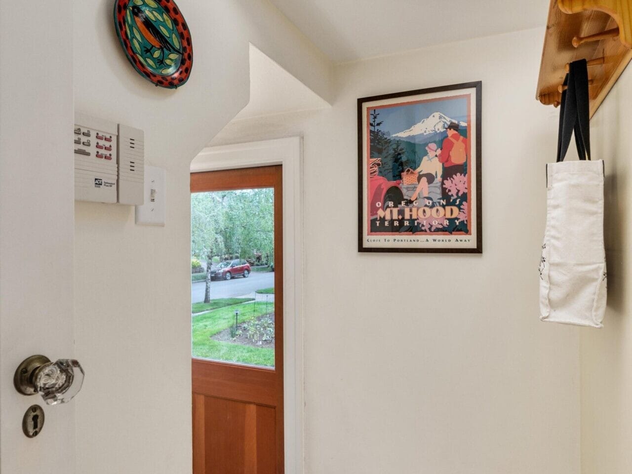 A small entryway with wooden steps leads to a doorway, inviting you into a glimpse of Portland, Oregon's lush greenery. A framed poster and decorated plate adorn the walls, while a tote bag hangs from the wooden coat rack.