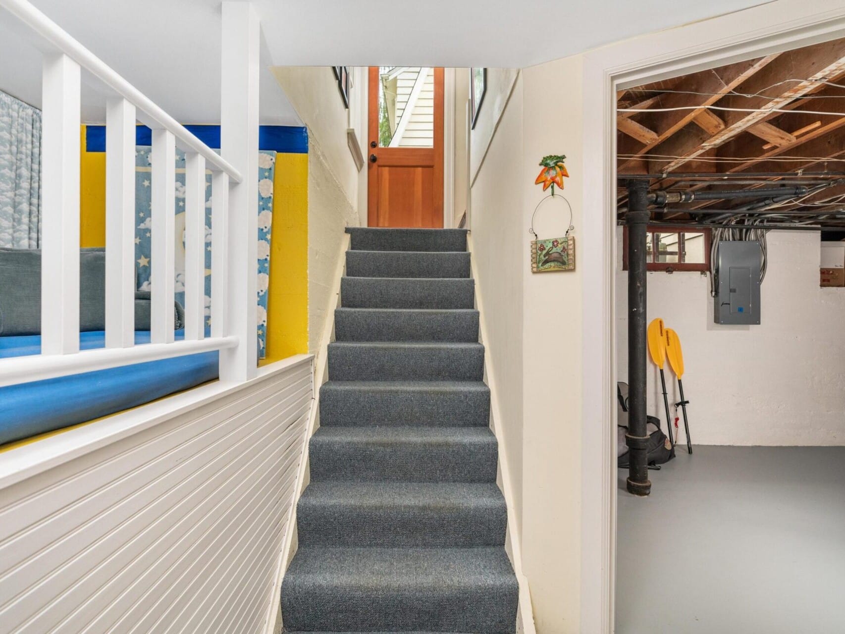 A carpeted stairway leads from a basement to the main floor in this Portland, Oregon home. On the right, the unfinished basement area features exposed beams and utility meters. A bright upper level beckons, with a wooden door partially visible at the top of the stairs.