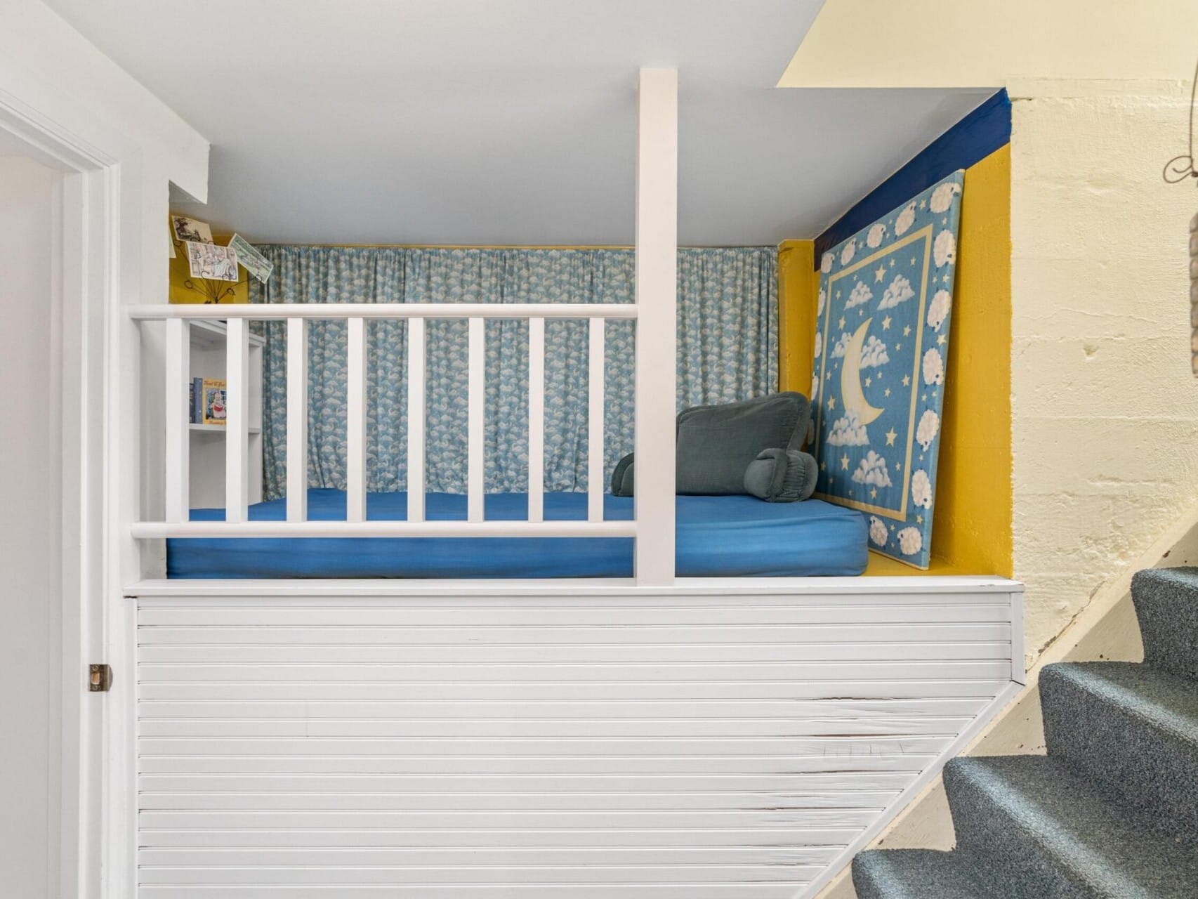 A cozy built-in reading nook in Portland, Oregon, features a white railing, blue cushion, and patterned pillows. The back wall is adorned with decorative fabric showcasing moon and star designs. Nestled beside a staircase with blue carpeting, it offers a tranquil escape in the heart of the city.