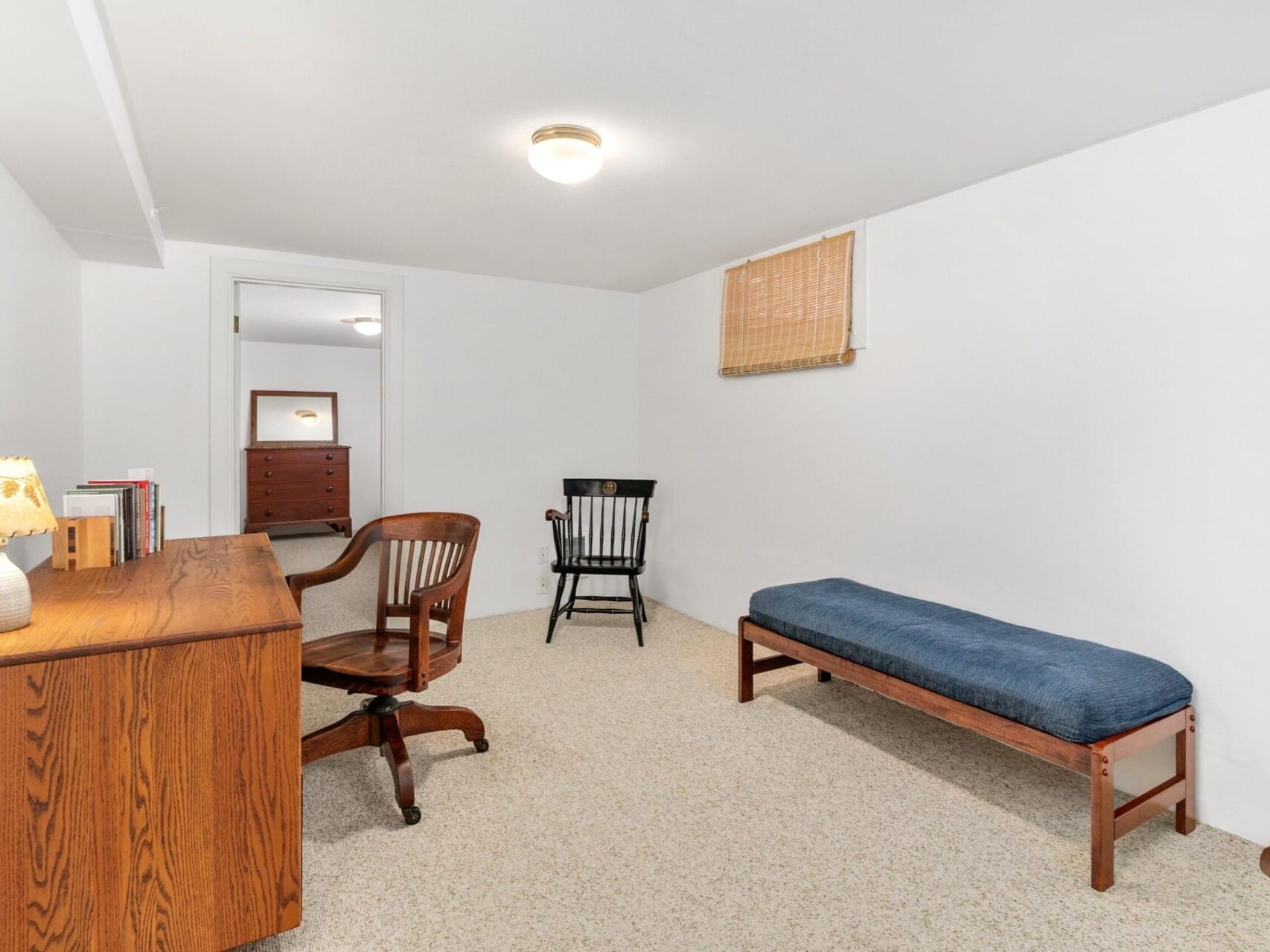 A small, minimalist room in Portland, Oregon boasts white walls and a wooden desk with a lamp and books. Two wooden chairs accompany a bench with a blue cushion, framed by a chest of drawers. Above the bench, a window features an elegant bamboo blind.