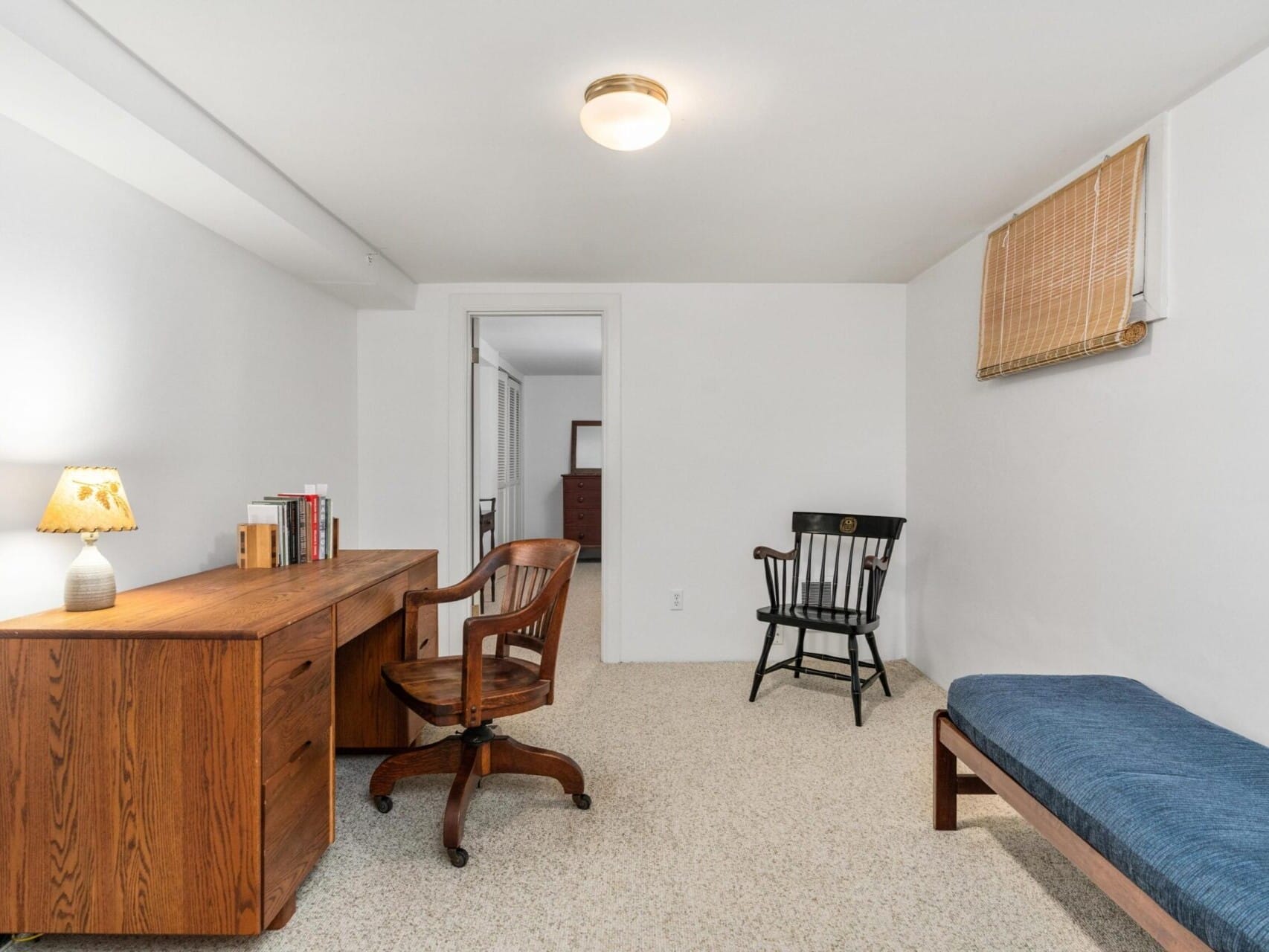 A minimalist room in Portland, Oregon, showcases white walls with a wooden desk holding books and a lamp, paired with a swivel chair. A black rocking chair and simple blue cushioned bench sit beneath a small window adorned with bamboo shades. The carpeted floor completes the cozy space.