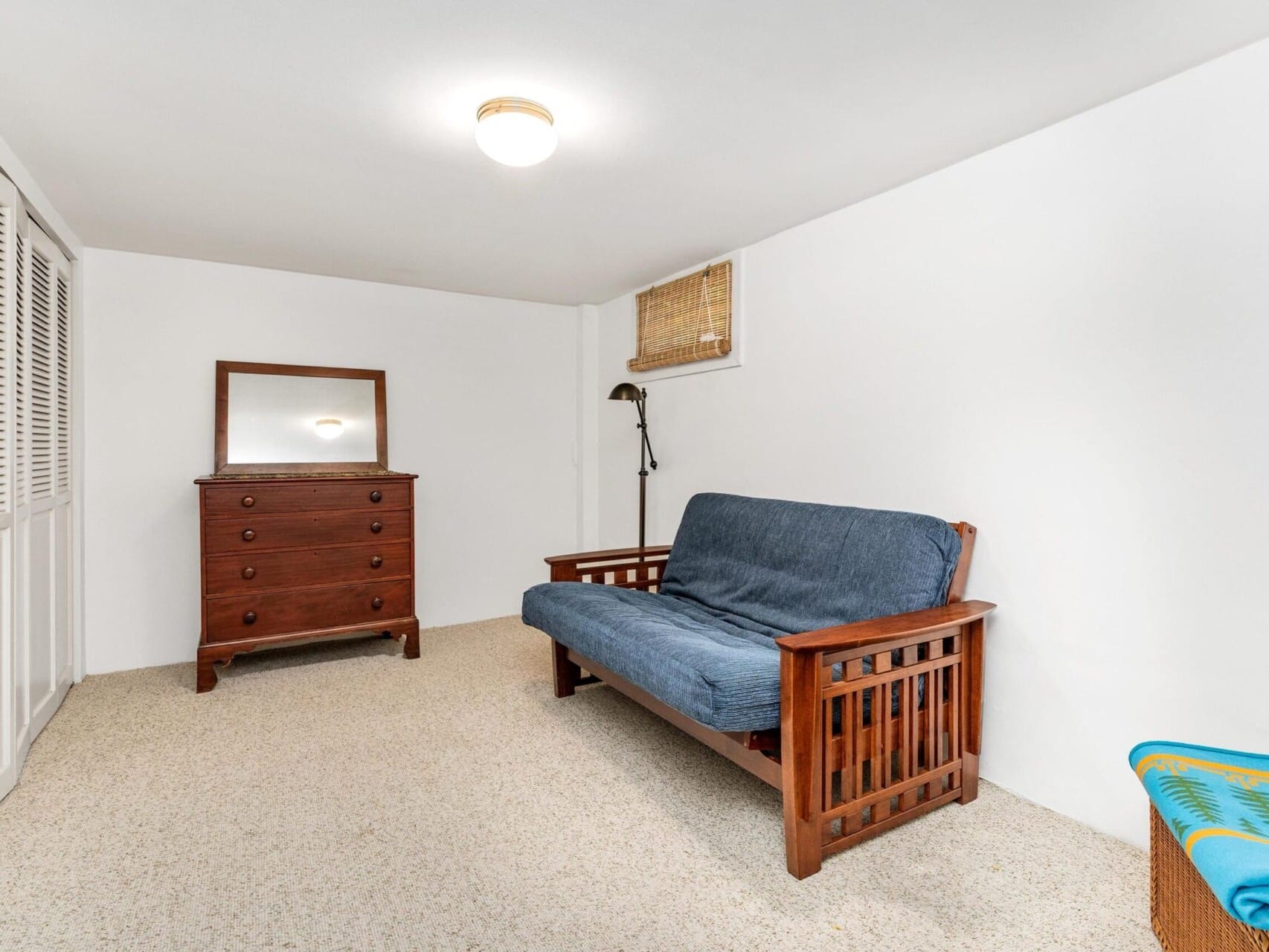 A simple room in Portland, Oregon, features a wooden dresser, a futon with a blue mattress, and a floor lamp. The walls are white, and there's a small window with a wicker shade. A wicker basket adorned with colorful cloth sits nearby.