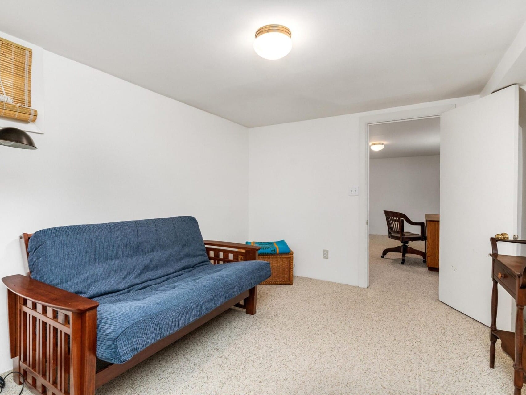 A small, well-lit room in Portland, Oregon, features a futon draped in a blue throw, a wooden desk and chair, and a closed door leading to another room. The plain white walls and light-colored carpet are gently illuminated by the ceiling-mounted light.