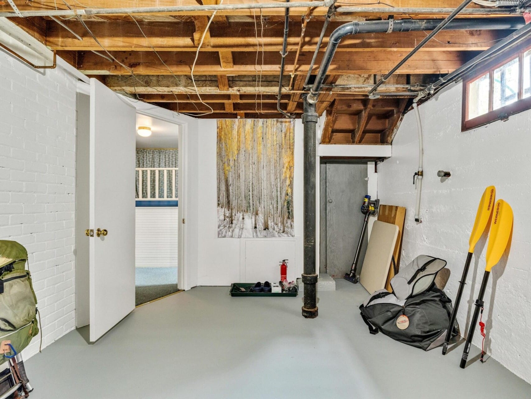 A basement in Portland, Oregon, features exposed wooden beams, white brick walls, and gray flooring. It contains a backpack, rolled-up items, two yellow paddles, and a framed forest artwork. An open door leads to a carpeted room with blue wallpaper and white trim.