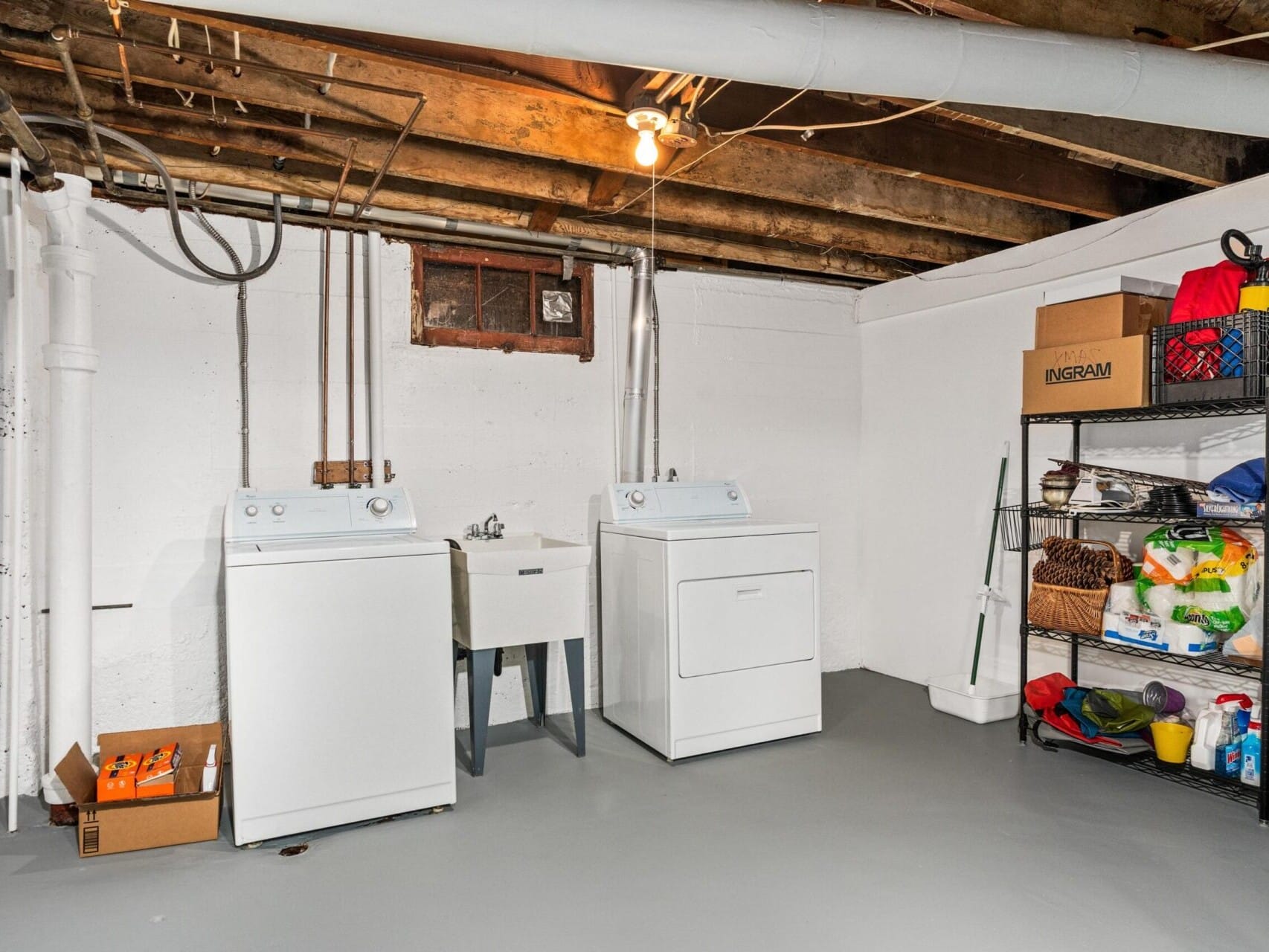 A basement laundry room in Portland, Oregon, features a washer, dryer, and utility sink. Exposed ceiling beams and a small window give it a rustic charm. On the right, metal shelving holds various household items and supplies.