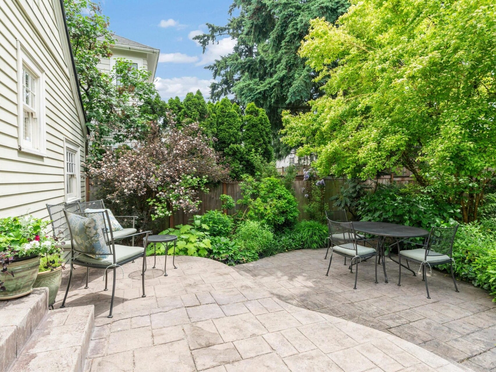 A cozy backyard patio in Portland, Oregon features metal chairs and tables surrounded by lush greenery and colorful plants. A light beige house wall is on the left, while stone tiles pave the inviting space beneath a clear blue sky.