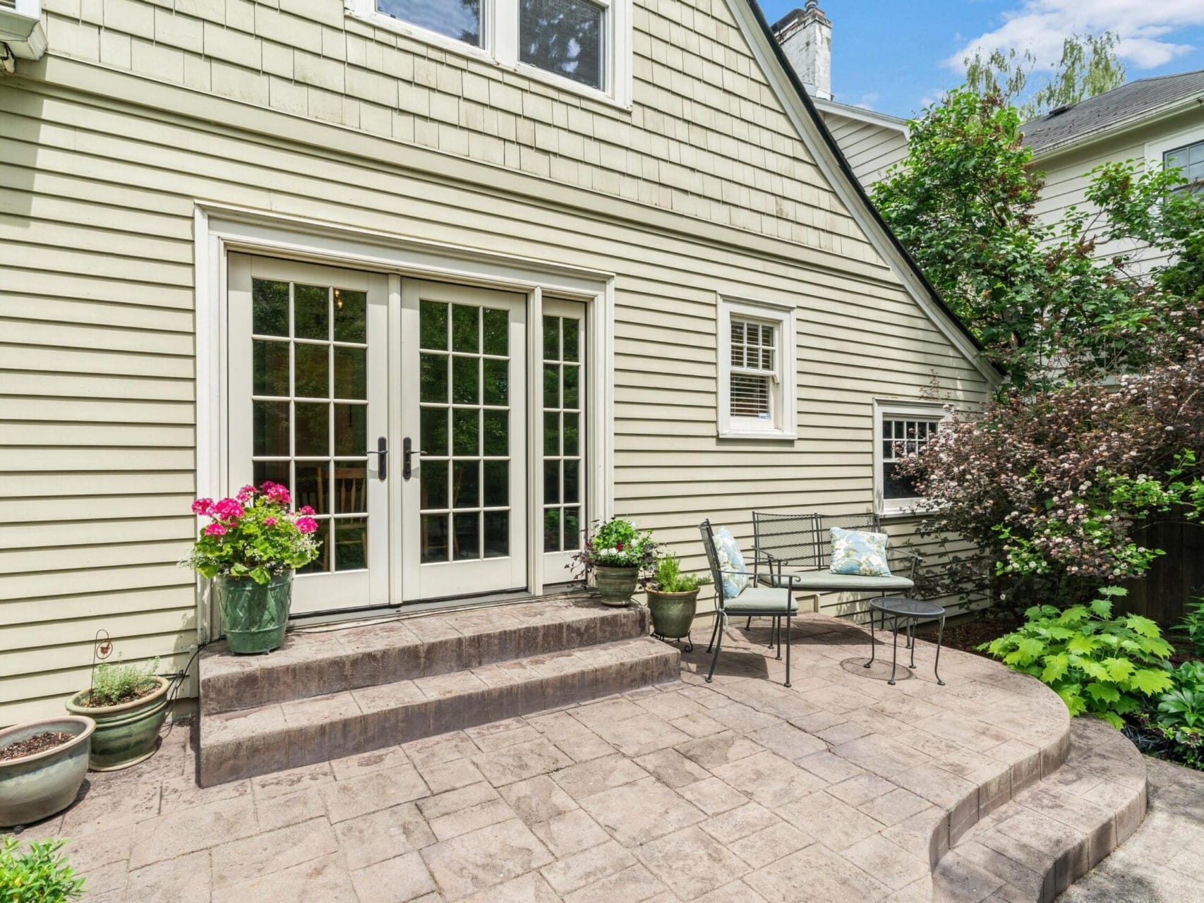 A cozy Portland, Oregon patio with stone flooring features two metal chairs and a small table, lush greenery on the side, and vibrant pink flowers in pots next to a set of glass-paneled French doors leading into a house with beige siding.