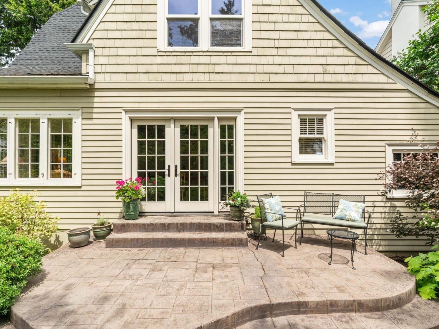 A charming house in Portland, Oregon, boasts light green siding and a sloped roof. It features French doors that open to a stone patio with two chairs and colorful potted flowers. Lush greenery envelops the area, complemented by large windows adorning the house.