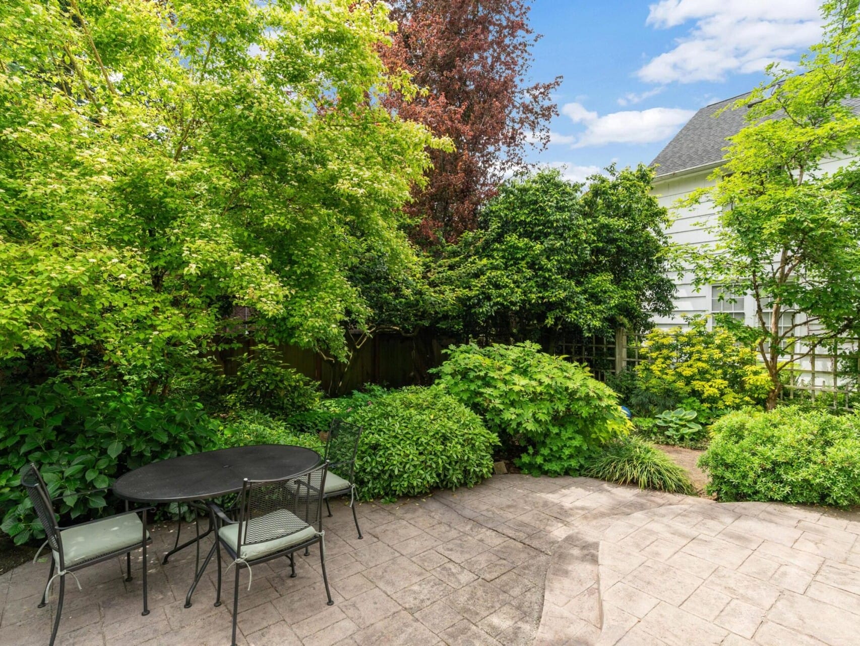 A tranquil Portland, Oregon backyard with a stone patio featuring a round metal table and two chairs. Lush green trees and bushes surround the patio, while a glimpse of a white house with a dark roof is visible in the background under a partly cloudy sky.