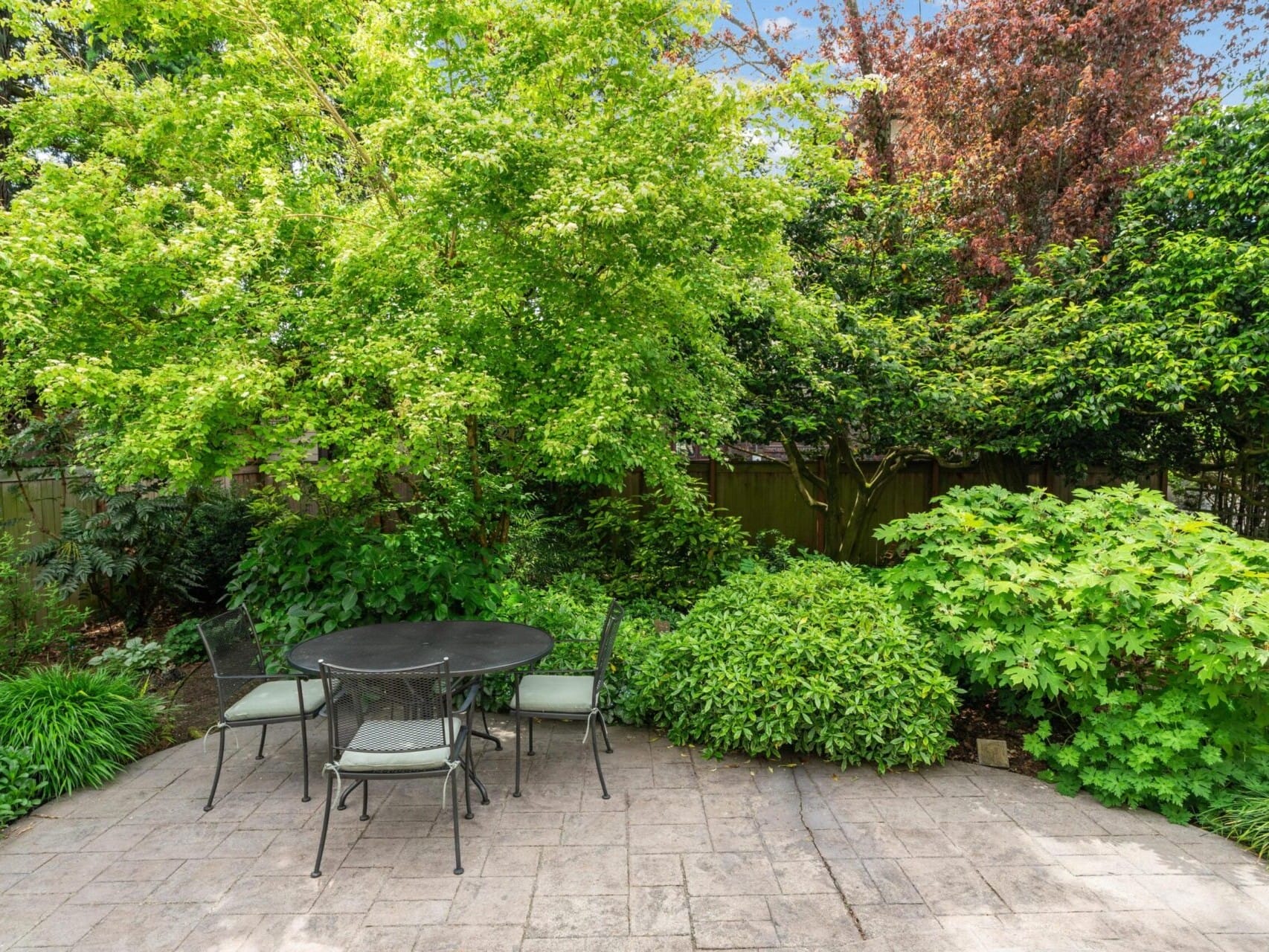 A peaceful backyard patio in Portland, Oregon, with a black metal table and four chairs sits amidst lush greenery, including trees and shrubs, under a clear blue sky.