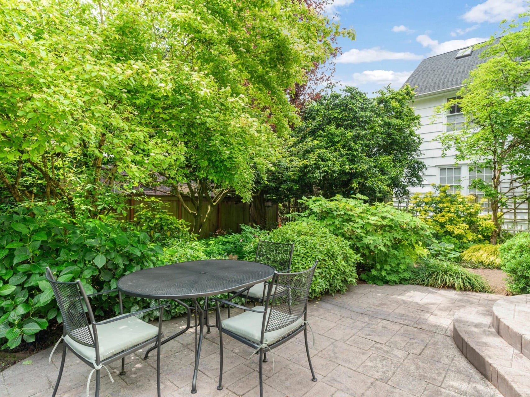 A serene Portland, Oregon backyard features a round black metal table with four chairs on a stone patio, surrounded by lush greenery and trees. In the background, a white multi-story house is partially visible under the clear blue sky.