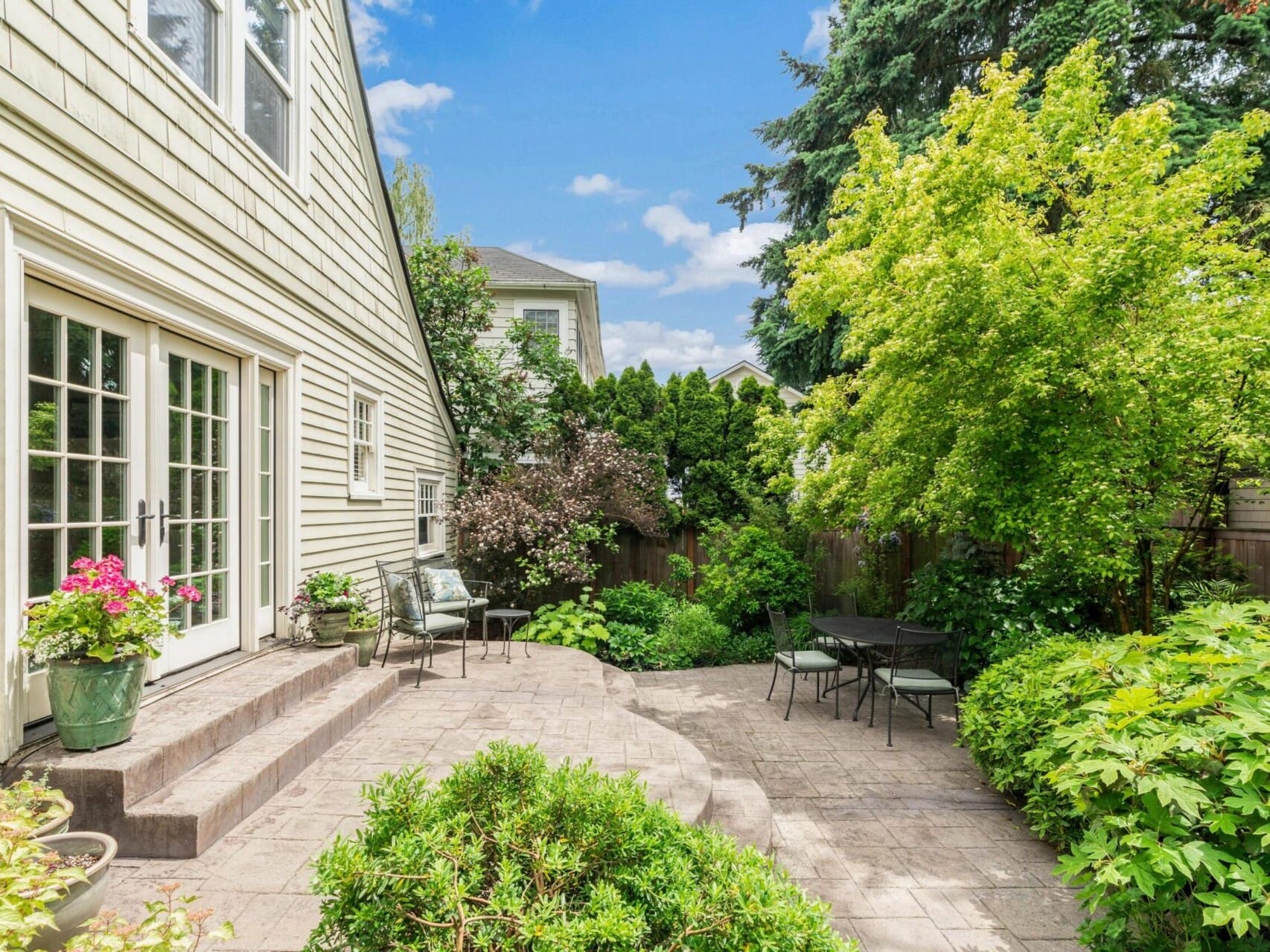 A quaint Portland, Oregon backyard features a paved patio area with a small table and chairs. Lush greenery, including trees and shrubs, surrounds the space, creating a serene atmosphere. French doors open from the house, adding charm to the garden setting.