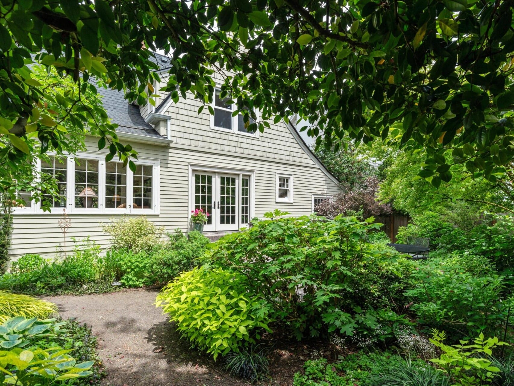 A charming house with white siding and numerous windows, nestled in the lush greenery of Portland, Oregon. A well-maintained garden path winds through dense trees and bushes, framing a tranquil, secluded atmosphere.