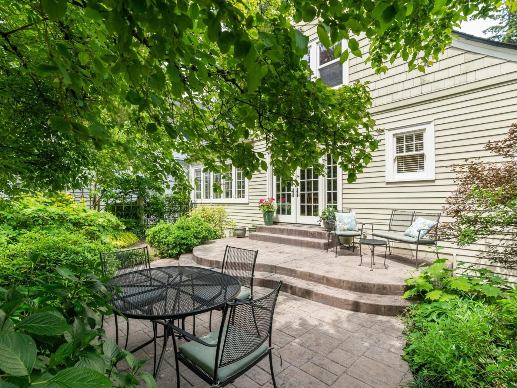 A cozy backyard patio in Portland, Oregon, features a round metal table with chairs, surrounded by lush greenery. Steps lead up to the light-colored house with large windows, creating a serene and inviting outdoor atmosphere.