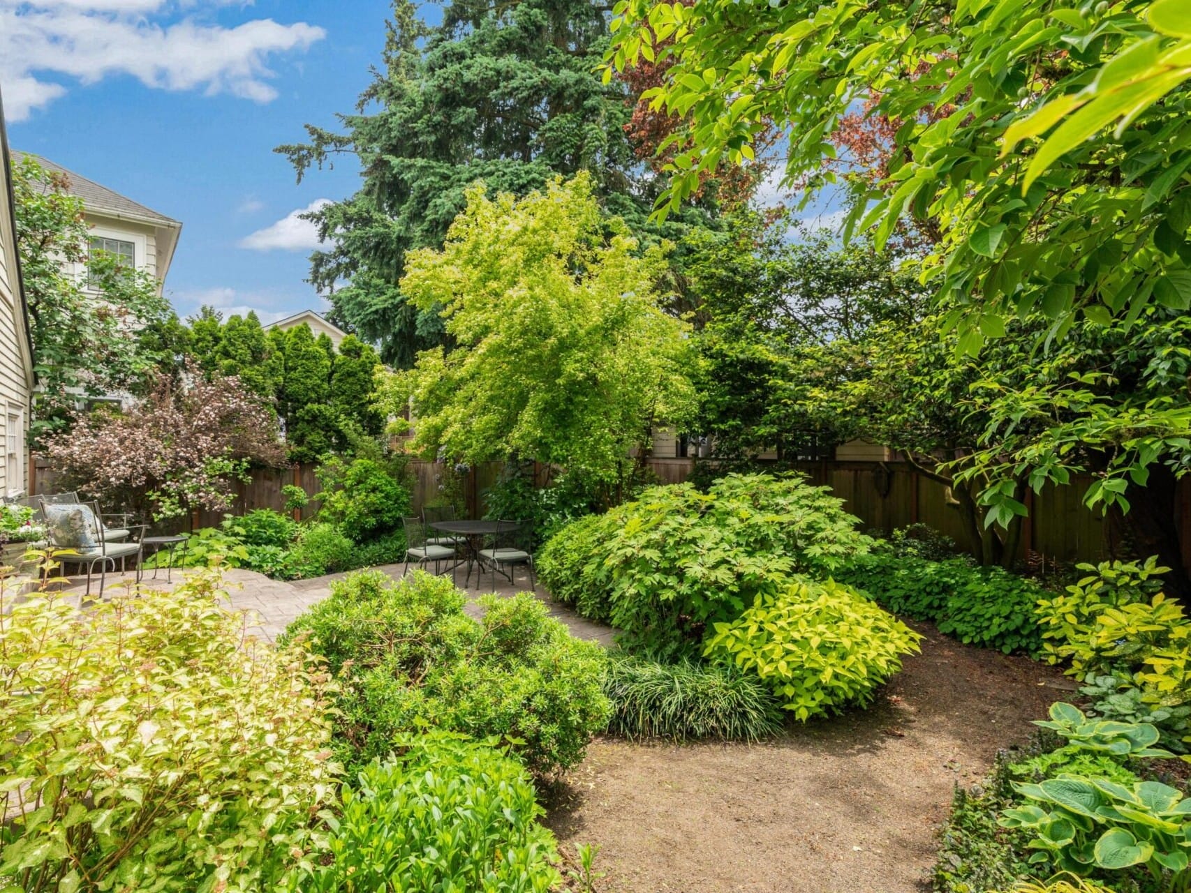 A lush garden in Portland, Oregon, boasts a variety of green shrubs and trees surrounding a small, circular table with two chairs. The space is bordered by wooden fencing, with a house partially visible on the left under the bright blue sky.