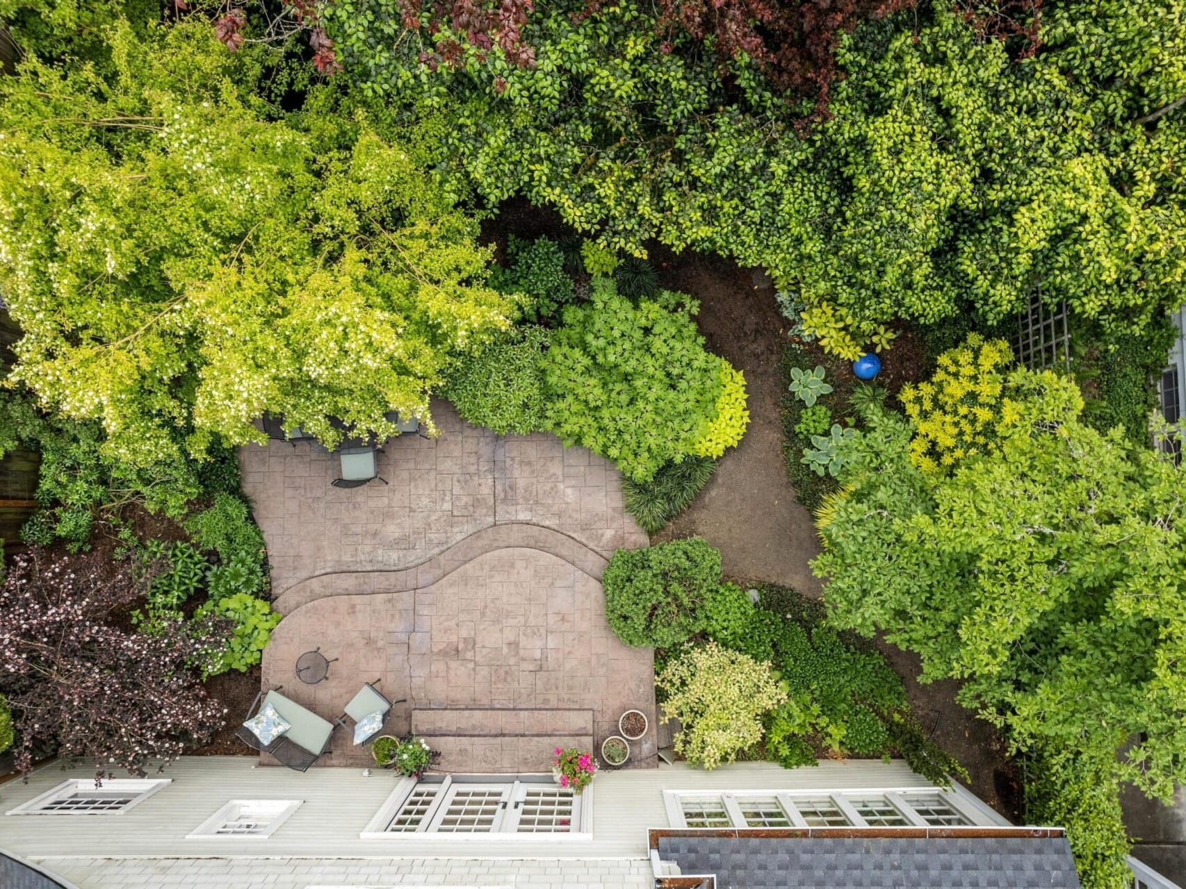 Aerial view of a garden in Portland, Oregon, with lush greenery surrounding a stone patio. The patio features outdoor furniture, including chairs and a table. A blue ball is visible in the garden area. The space is beautifully framed by the dense trees and shrubs typical of this region.