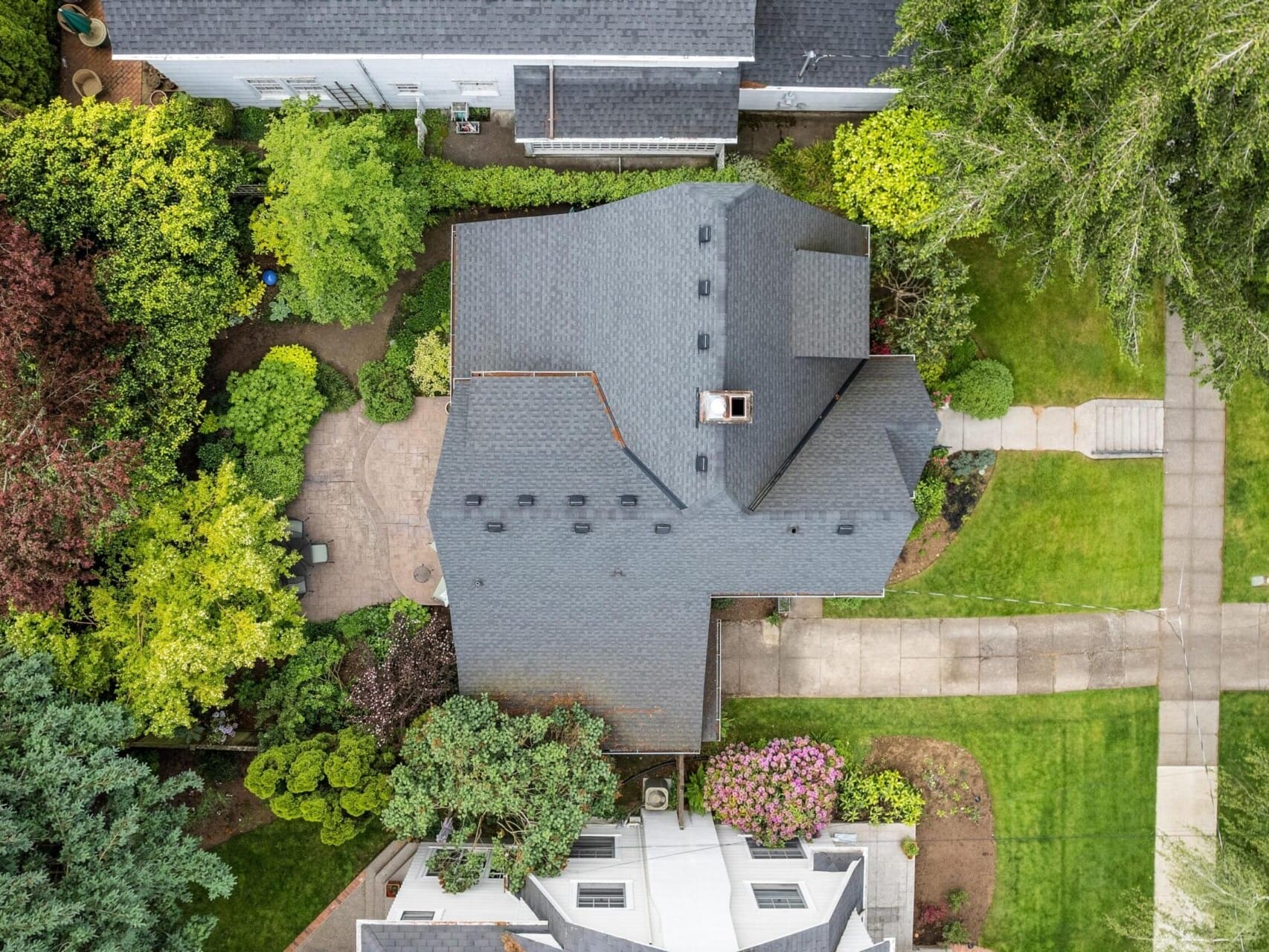 Aerial view of a house with a dark roof in Portland, Oregon, surrounded by lush green trees and neatly trimmed lawns. A stone pathway leads to the entrance, and a concrete sidewalk runs parallel to the street.