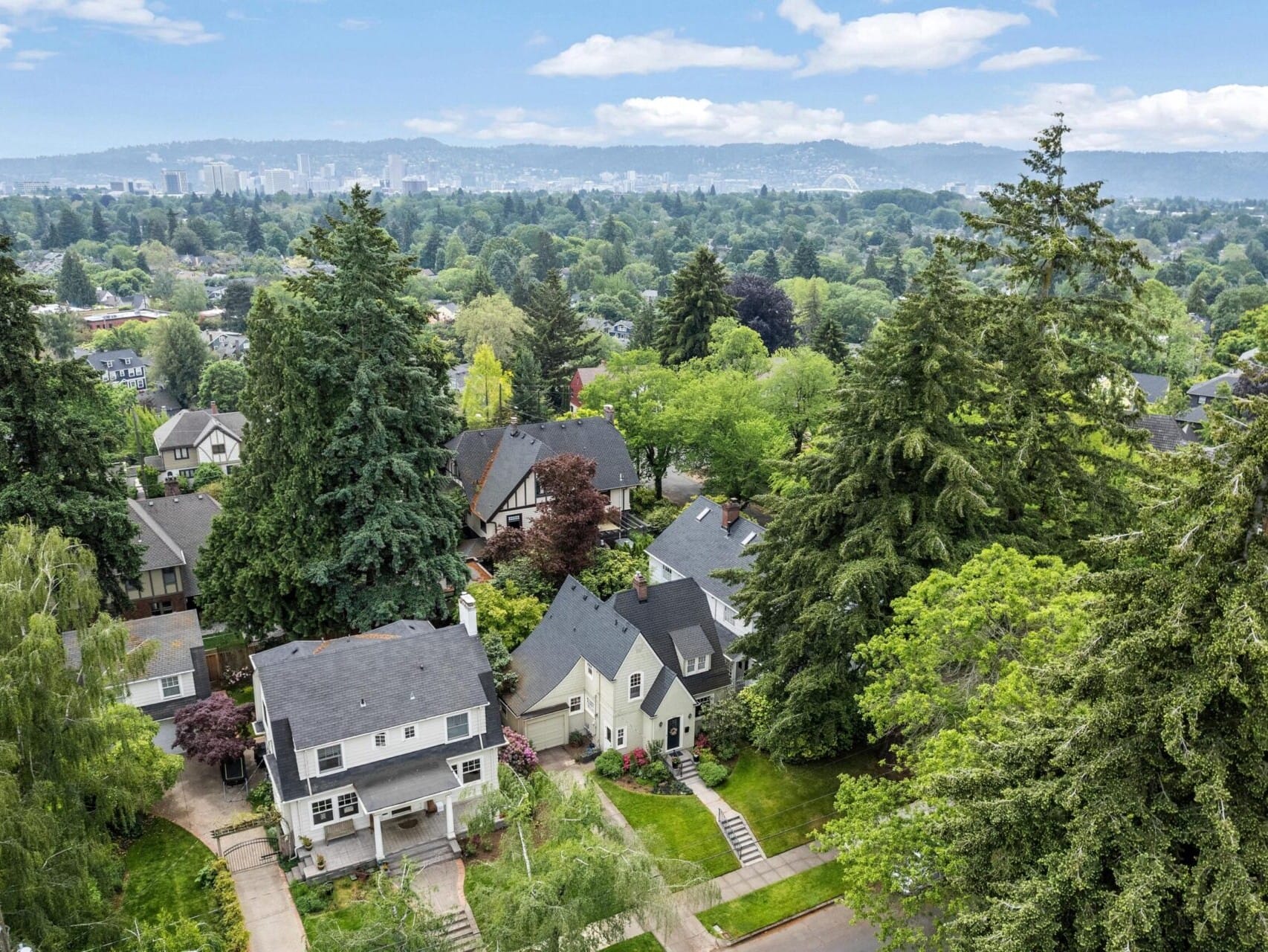 Aerial view of a suburban neighborhood in Portland, Oregon, with houses surrounded by tall trees. The city skyline is visible in the distance under a partly cloudy sky. Lush greenery dominates the scene, creating a serene and picturesque atmosphere.