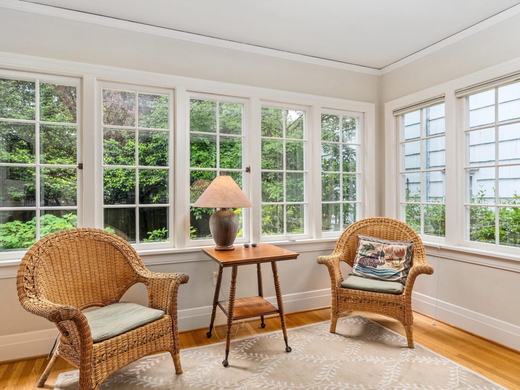 A bright sunroom in Portland, Oregon, featuring two wicker chairs with cushions, a wooden side table with a lamp, and large windows showcasing a lush green garden. The wooden floor is partially covered by a patterned rug.