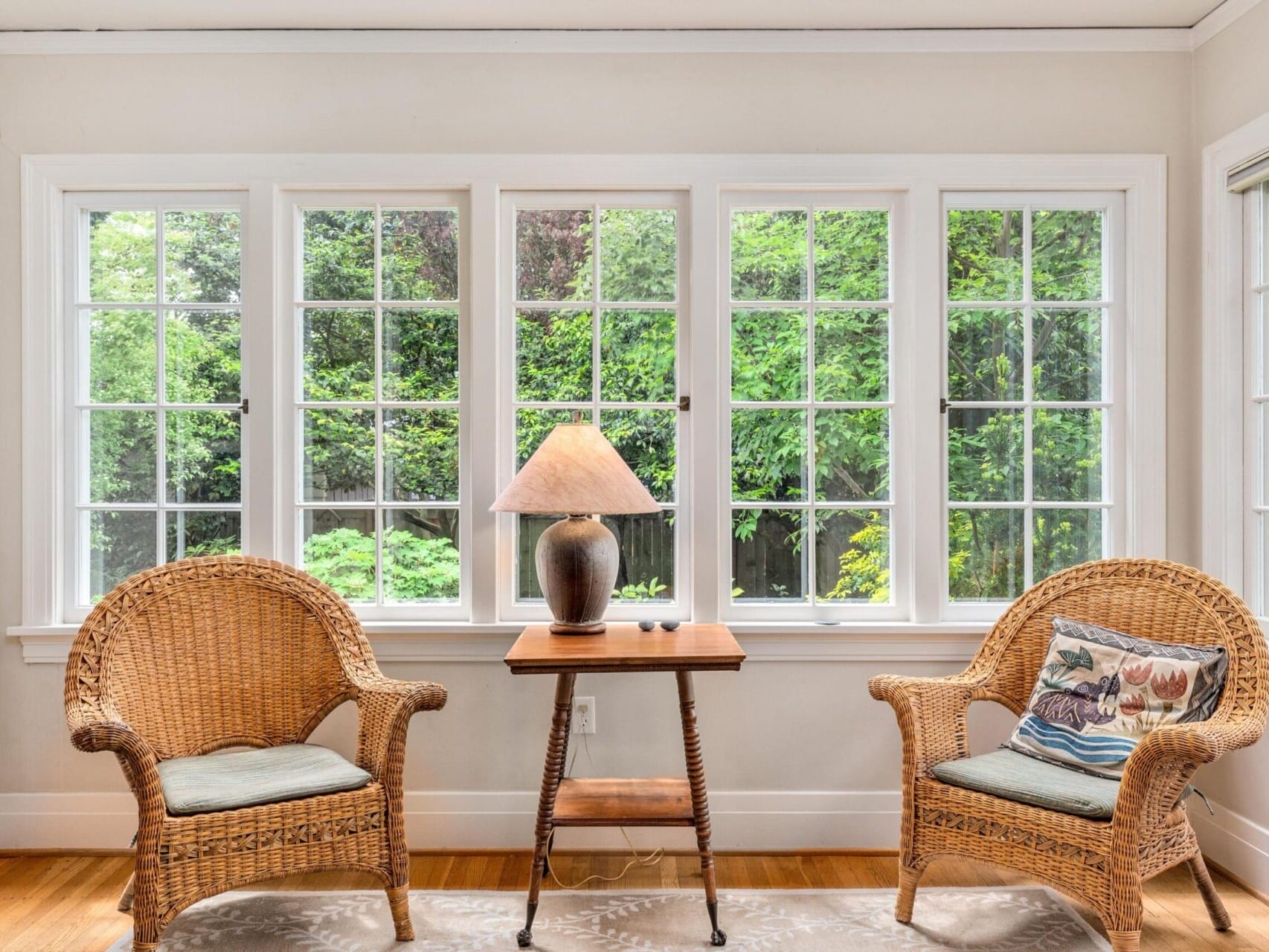 A cozy sunroom in Portland, Oregon, features two wicker chairs facing a wooden table with a lamp. Large windows reveal the lush green garden outside. A patterned rug and decorative cushions add warmth to this inviting space.