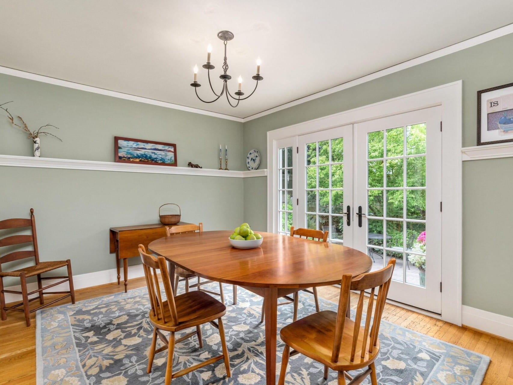 A dining room in Portland, Oregon, boasts green walls and a round wooden table with four chairs. A bowl of green apples sits on the table. There’s a floral-patterned rug, a wooden side table, and a chandelier above. French doors open to the lush garden outside.