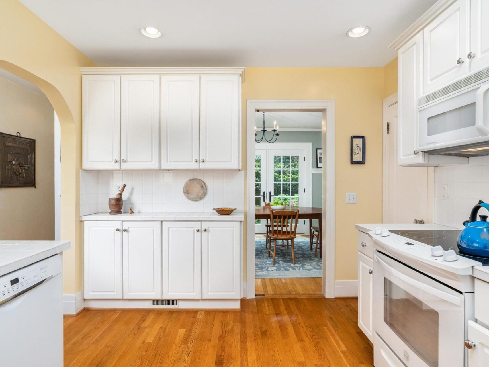 A bright kitchen in Portland, Oregon features white cabinets and wood flooring. A blue kettle sits on the stove. Through an open doorway, a dining area with a wooden table and chairs is visible, accompanied by a window and a chandelier.