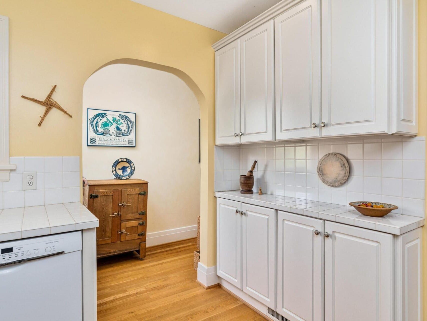 A Portland, Oregon kitchen showcases white cabinets and a dishwasher on the left. An archway guides you to a room with a wooden dresser and decorative wall art. The yellow walls add warmth, complemented by a small display bowl on the counter and a wheel adorning one wall.