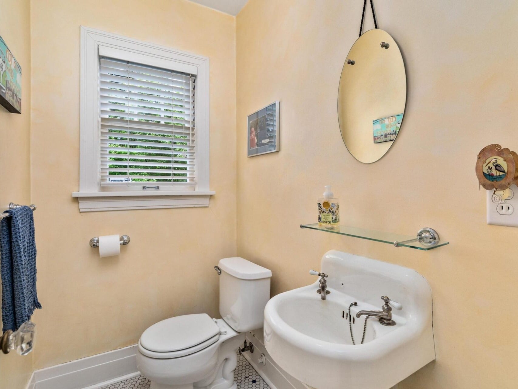 A small bathroom in Portland, Oregon, with light yellow walls features a white toilet and a vintage-style sink with two faucets. Above the sink is an oval mirror and a glass shelf with soap. A window with blinds is above the toilet, and a dark towel hangs on the left.