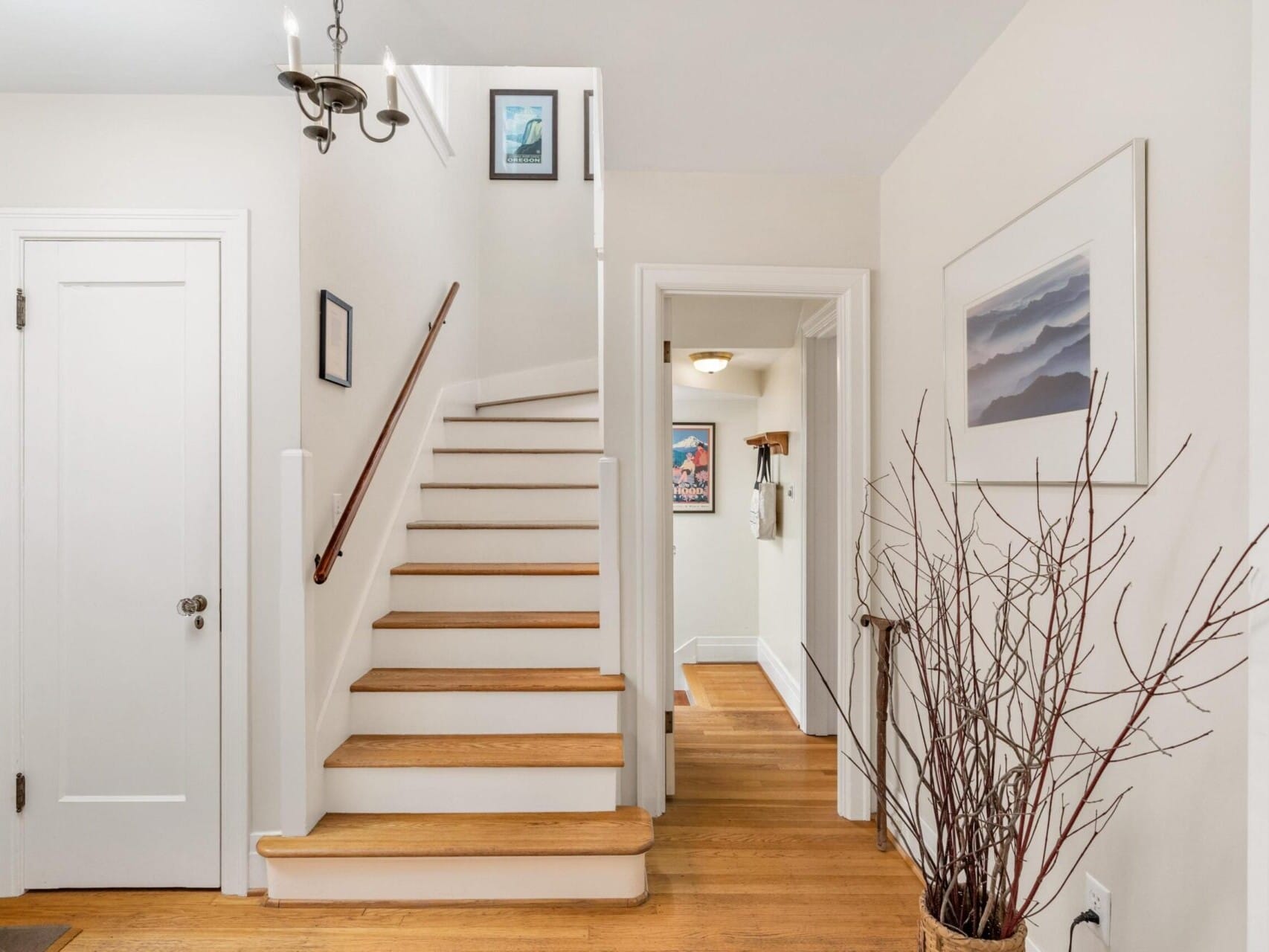 A bright hallway in Portland, Oregon, boasts wooden flooring and a staircase leading upwards. The walls are adorned with framed artwork, while a chandelier hangs elegantly from the ceiling. To the right, a vase with branches adds a touch of nature to this charming space.