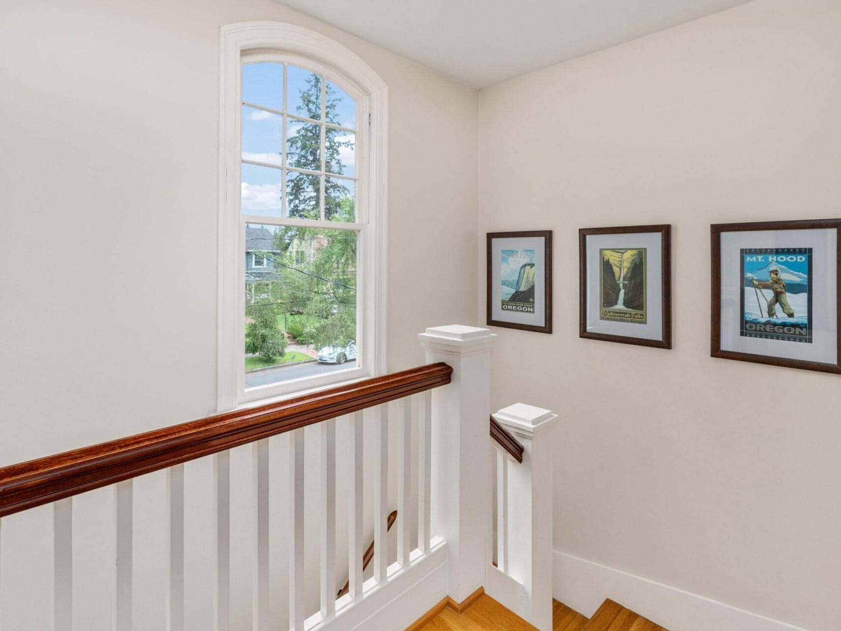 A bright hallway with a wooden handrail leads to a window overlooking a charming Portland, Oregon neighborhood. Three framed posters featuring nature scenes adorn the white walls, while sunlight streams in, creating a warm and inviting atmosphere.