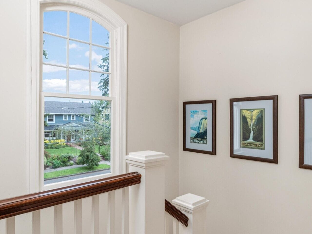 A bright staircase landing with white walls and wooden floors in a Portland, Oregon home. Three framed pictures grace the wall, while a large arched window bathes the space in natural light, offering a serene view of greenery and a distant house.