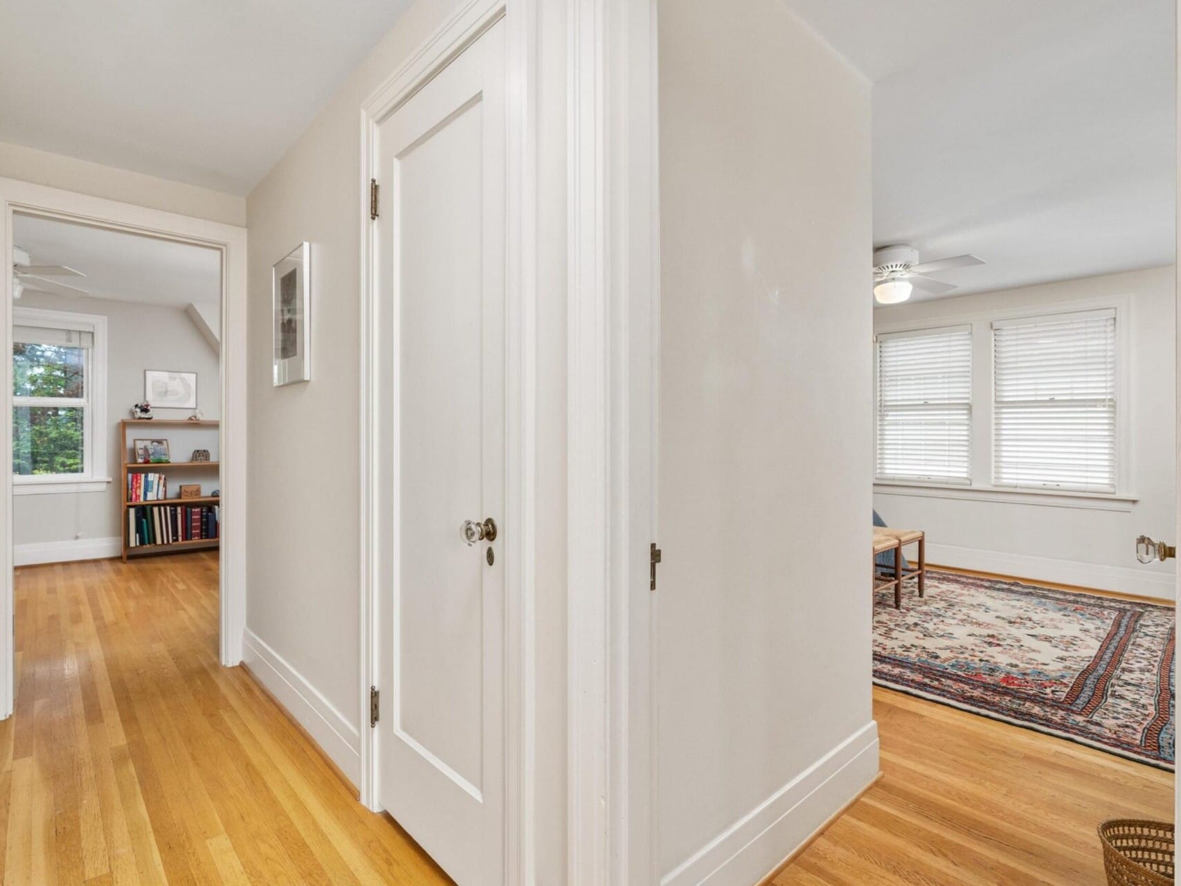 A hallway with light wooden floors and white walls greets you in this Portland, Oregon home. To the right, an entrance leads to a room featuring a colorful area rug and windows with blinds. The hallway extends to another room adorned with a bookshelf and a window view of lush greenery.