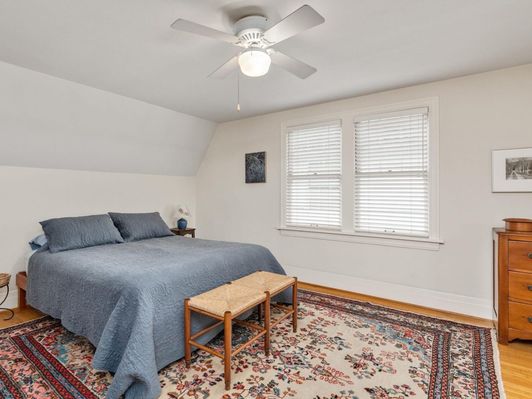 A Portland-inspired bedroom features a large bed with a blue bedspread, a wooden bench at the foot, and two nightstands. A ceiling fan whirs softly above a colorful patterned rug. The room has white walls, a window with blinds, and framed art reminiscent of Oregon's scenic beauty adorning the walls.