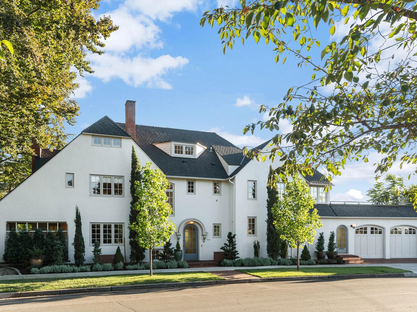 A large, elegant white house in Portland, Oregon with a dark roof features multiple windows and a central arched entrance. The property boasts neatly trimmed bushes and trees in the front yard and a driveway leading to a garage beneath the clear, sunny sky.