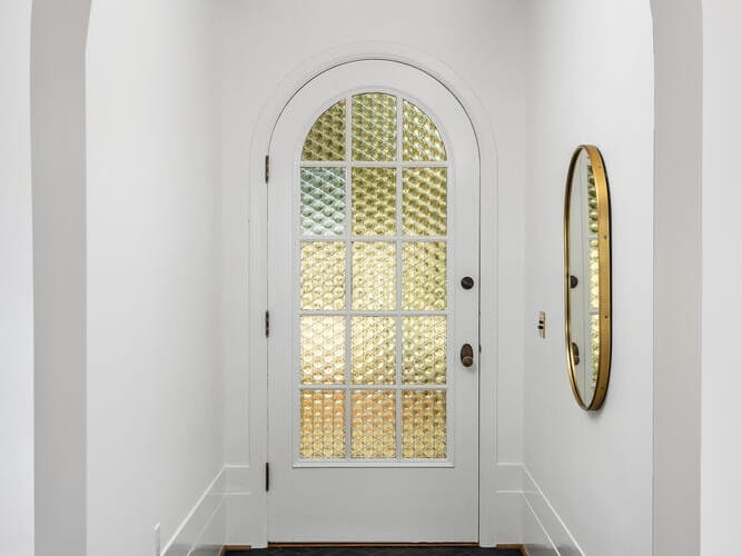 A hallway in Portland, Oregon, with white walls and a black herringbone tile floor leads to a door featuring a large arched glass panel. A round mirror graces the right wall, while a small ceiling light fixture is perched above the door.