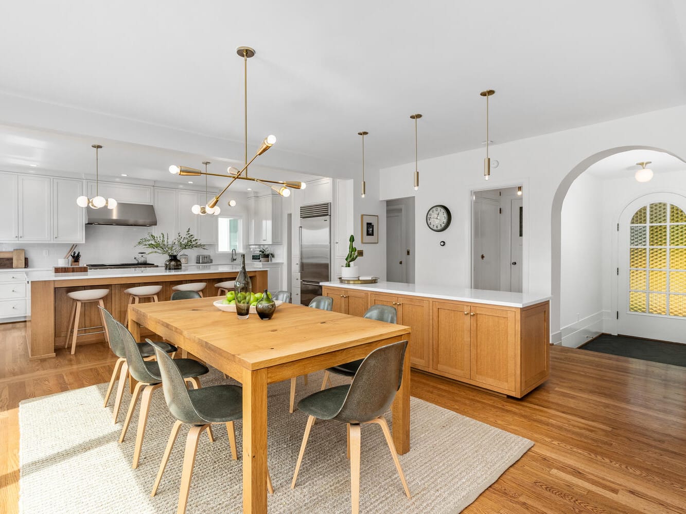 A modern kitchen and dining area in Portland, Oregon features a wooden table with green chairs and a stylish light fixture above. The kitchen boasts white cabinets, stainless steel appliances, and a large island with bar stools, all set on elegant hardwood floors.