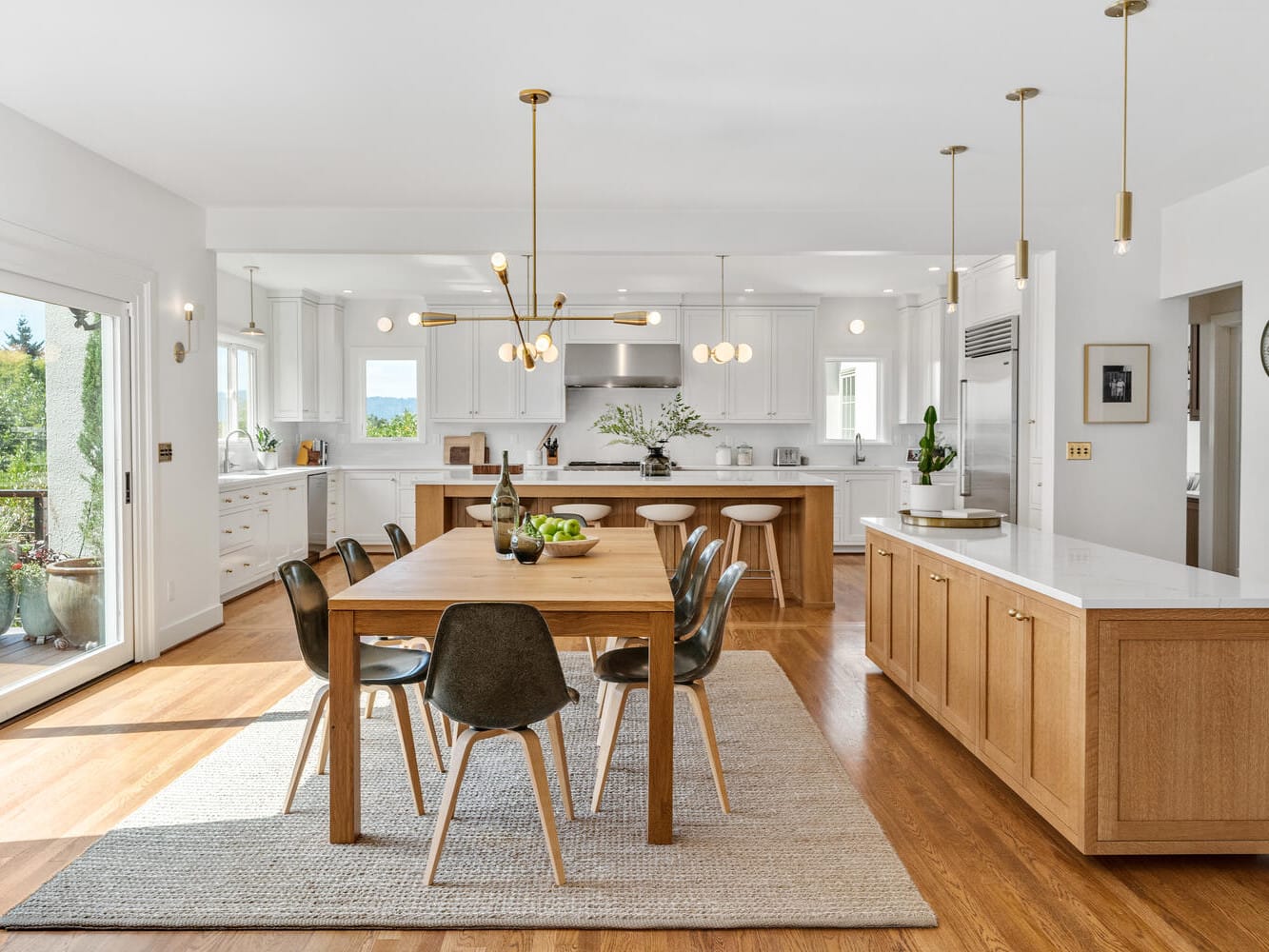 Spacious modern kitchen and dining area in Portland, Oregon, featuring a wooden table with black chairs on a rug, an island with light wood cabinets, white countertops, and multiple pendant lights. Large windows and glass doors provide natural light and garden views.