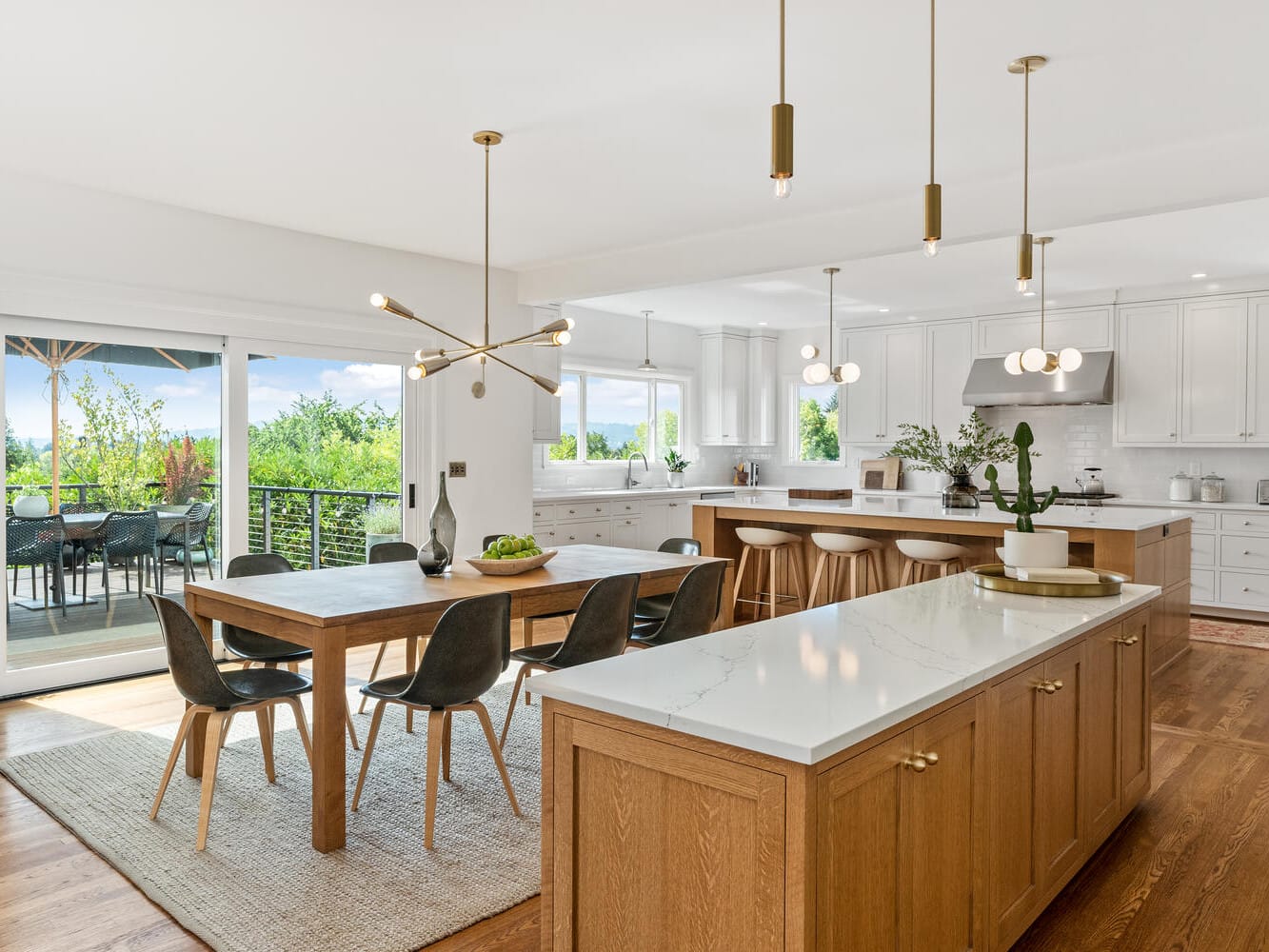 A modern kitchen in Portland, Oregon, features a wooden dining table with black chairs. The space boasts two islands with marble countertops, pendant lighting, and stainless steel appliances. Large windows and sliding doors open to a patio adorned with lush greenery outside.