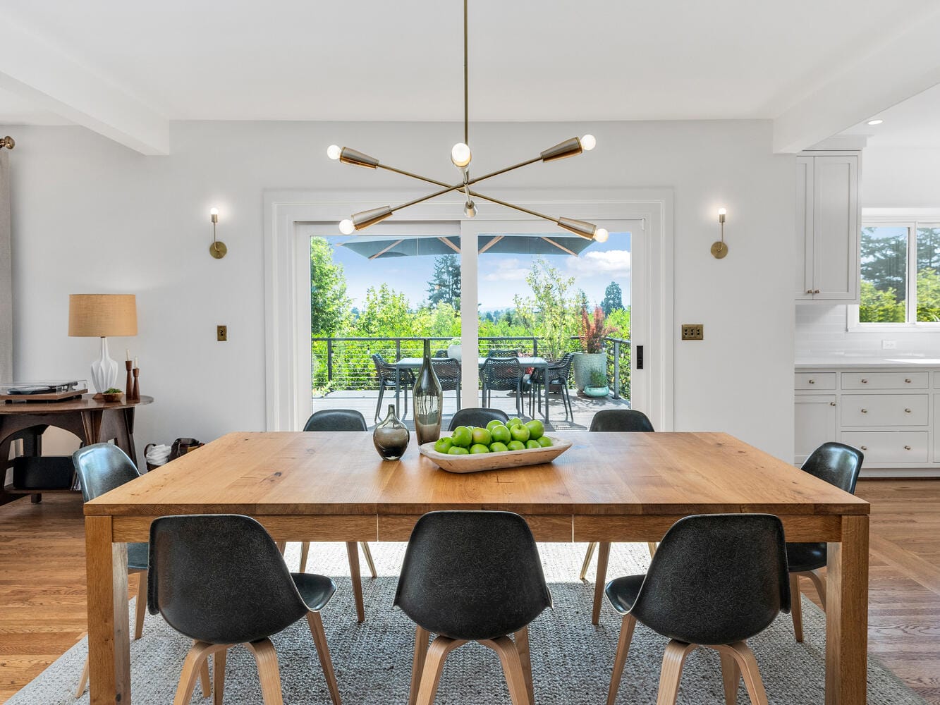 A modern dining room in Portland, Oregon features a wooden table with black chairs, a central fruit bowl with green apples, and a contemporary chandelier. Glass doors open to a view of the deck and lush greenery outside, creating a bright and inviting space.