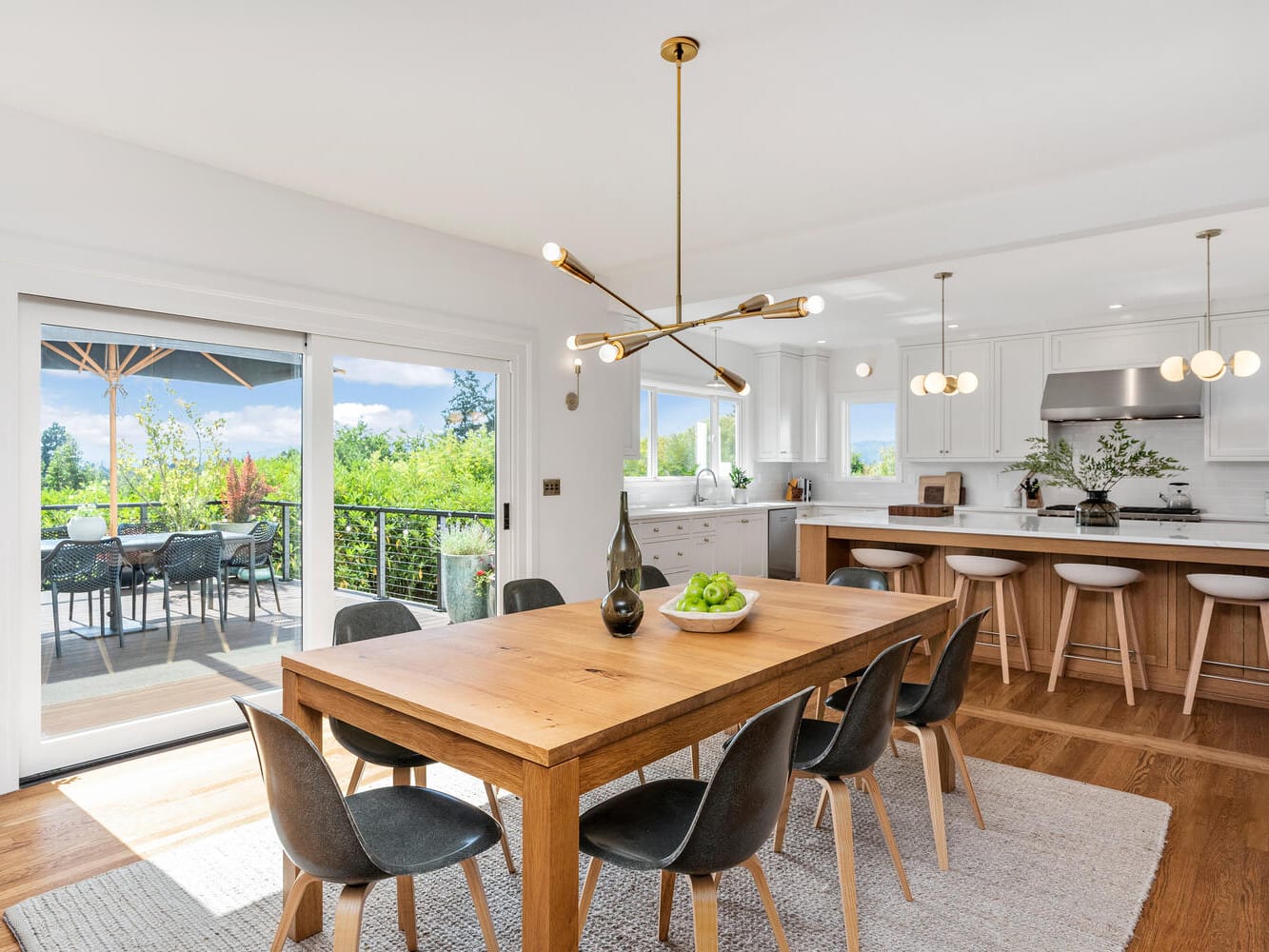 Spacious modern kitchen and dining area in Portland, Oregon, featuring a wooden table and black chairs. It boasts a large island, contemporary light fixtures, and sleek white cabinets. Sliding doors open to a patio with outdoor seating and a greenery view, filling the room with natural light.