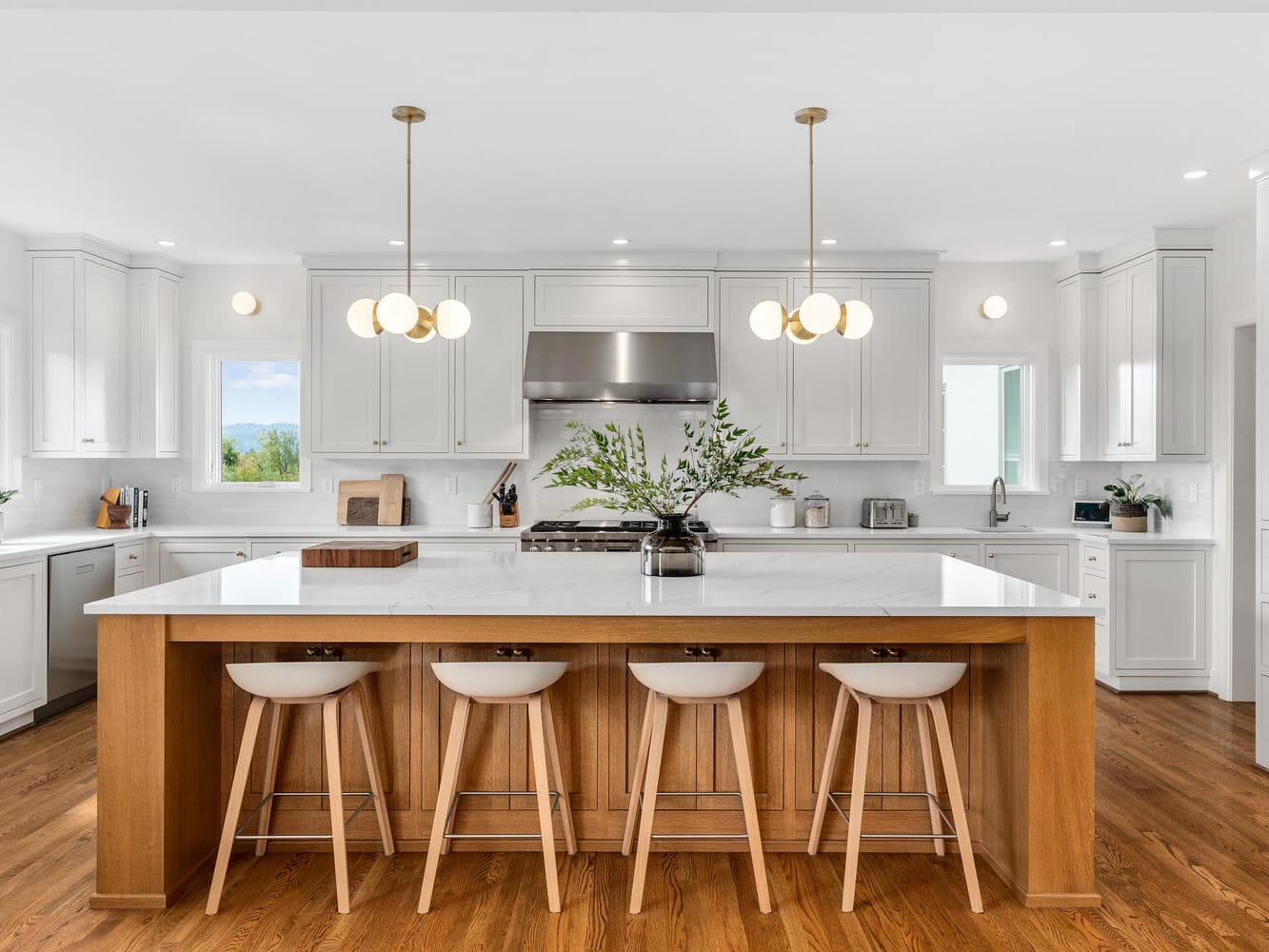 A modern kitchen in Portland, Oregon, features white cabinets and a large wooden island with four stools. Two pendant lights hang above the island, showcasing stainless steel appliances and a vase with green foliage. Wood flooring beautifully complements the bright space.