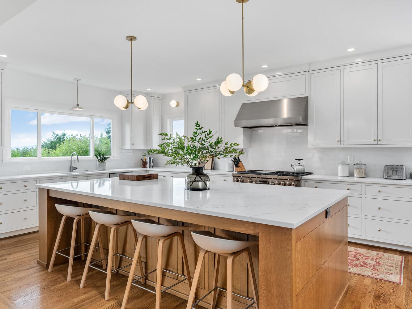 A modern kitchen in Portland, Oregon, featuring a large island with wooden stools, pendant lights, and a vase with greenery. White cabinets and a stainless steel stove are set against a backdrop of windows, providing natural light. Hardwood floors complete the space.